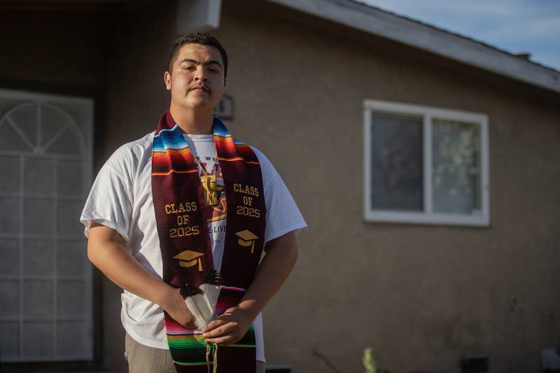 A man stands in front of a house wearing a colorful graduation stole.