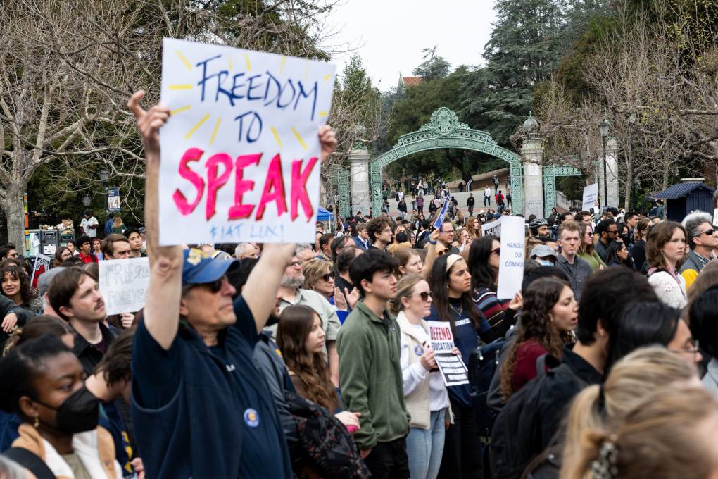 A student holds a sign saying 'Freedom to Speak' with several people standing in the background.