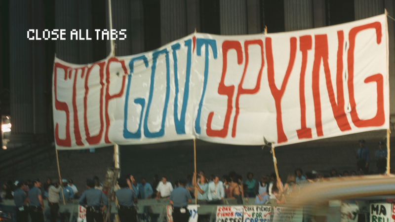 A group of people holding up a banner that says “Stop Government Spying” while police stand around them. The group of people appear to be protesting government surveillance and are blurry in the photo. So are the police.