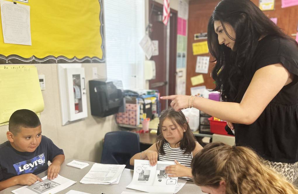 A woman wearing a sleeveless black shirt, points in the direction of a child who is sitting at a desk next to two other children in a classroom.