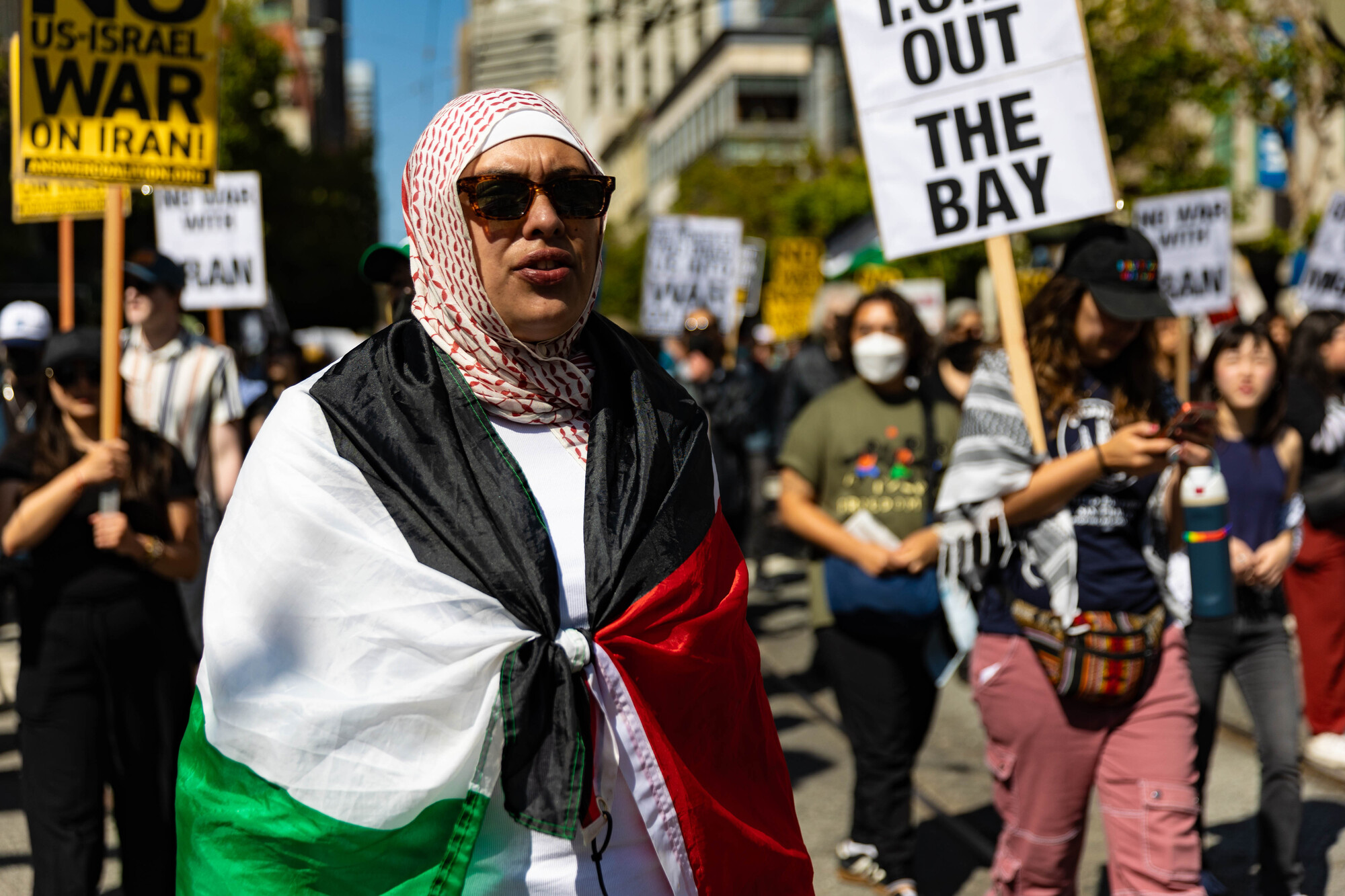 A woman wearing a red, white, green and black flag over her shoulders with sun glasses walks in a large group of people holding signs.
