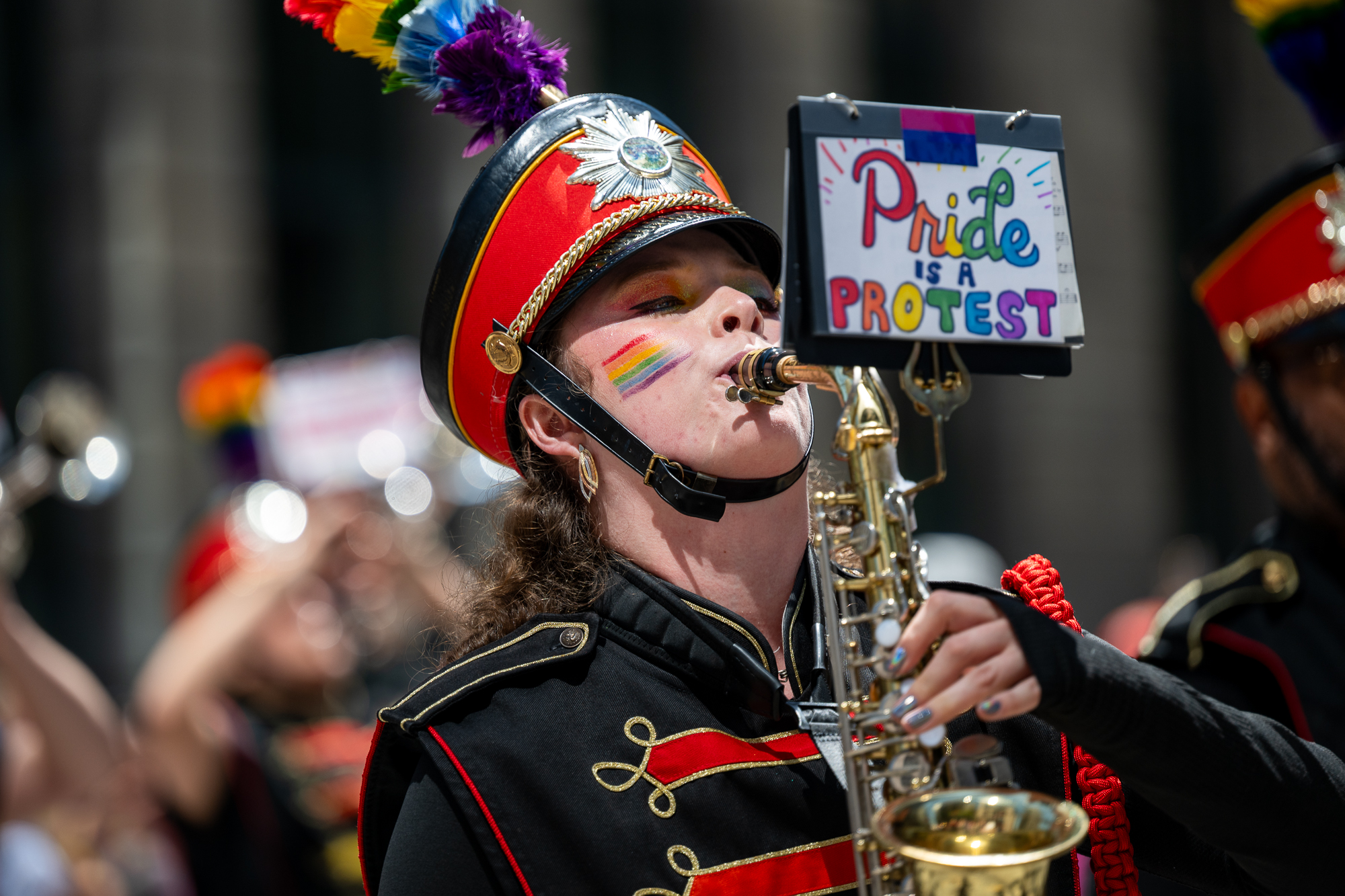 A person wearing a band outfit plays a saxophone with a sign that says Pride.