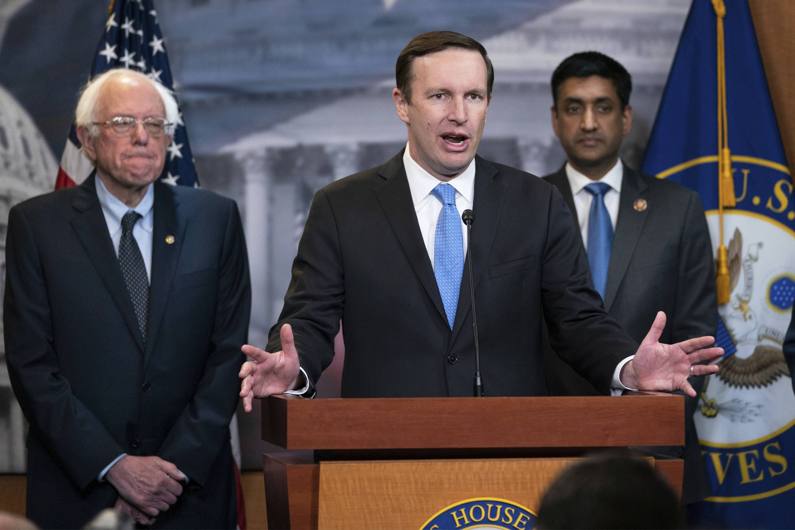 A man with a blue tie stands in the center at a podium with two other men in suits standing behind him.