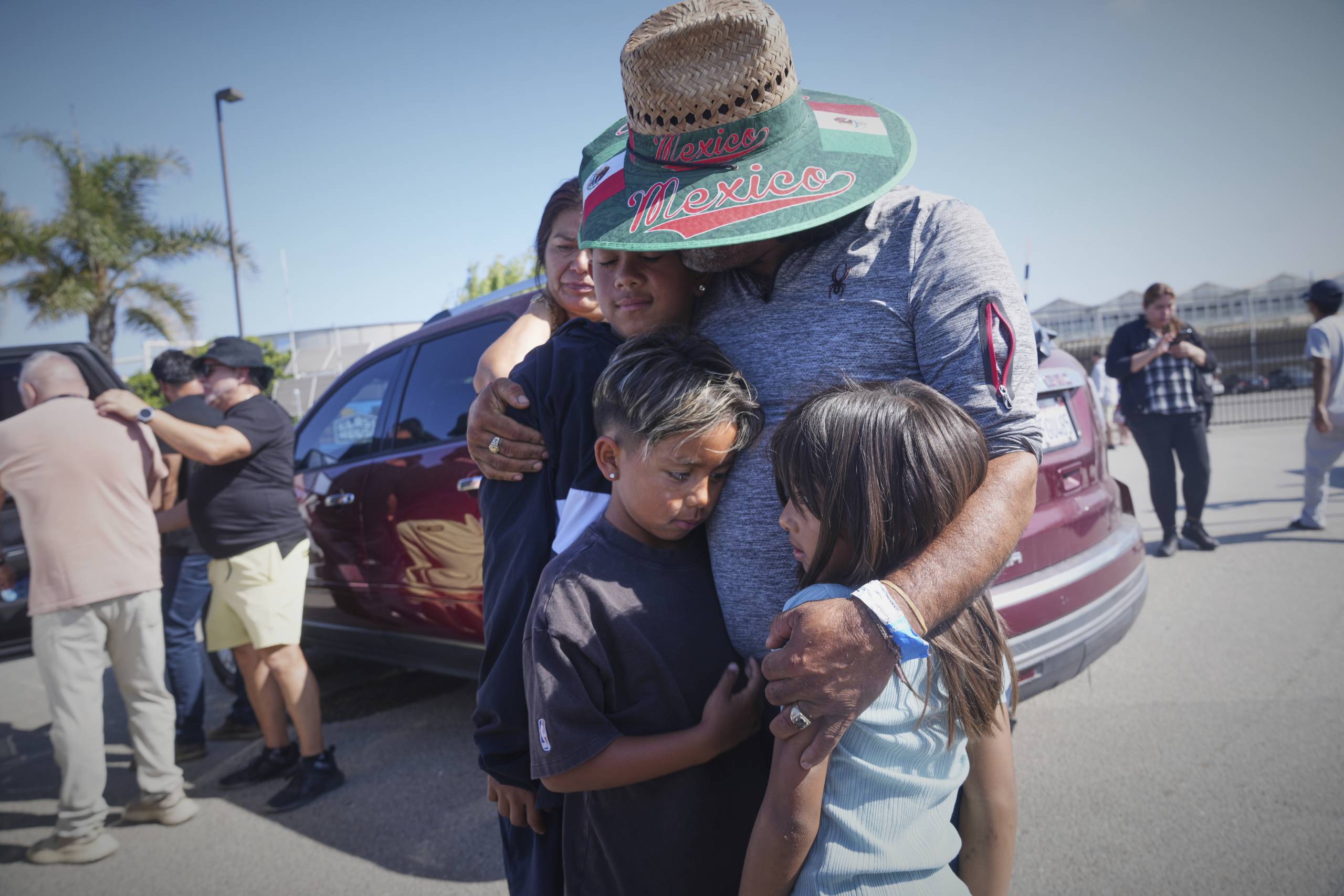 A man wearing a white-brimmed hat that says "Mexico" embraces three children and a woman outside in front of a red van.