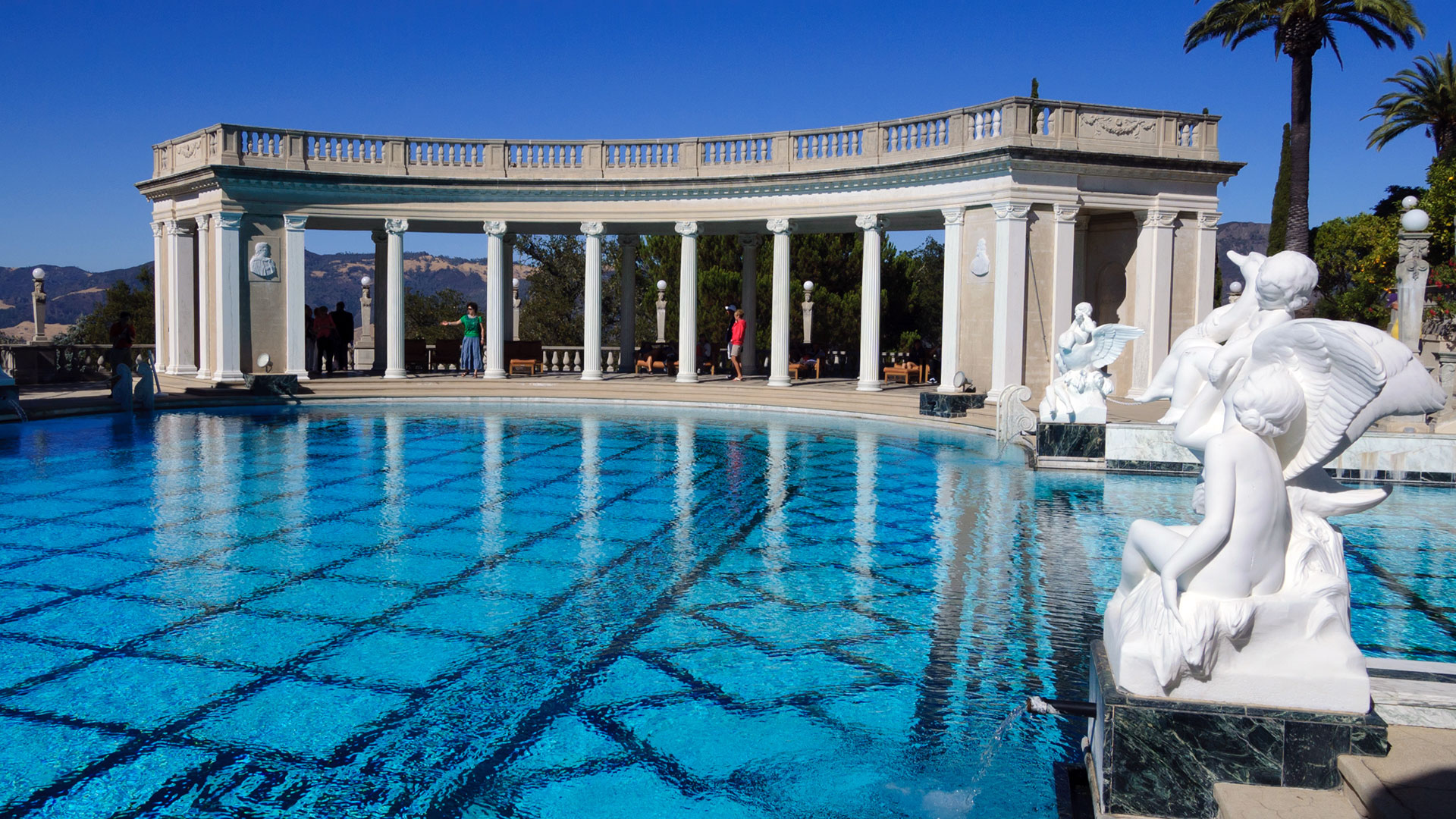 A brilliant aquamarine pool leads up to a stately building with arches and sculptures.