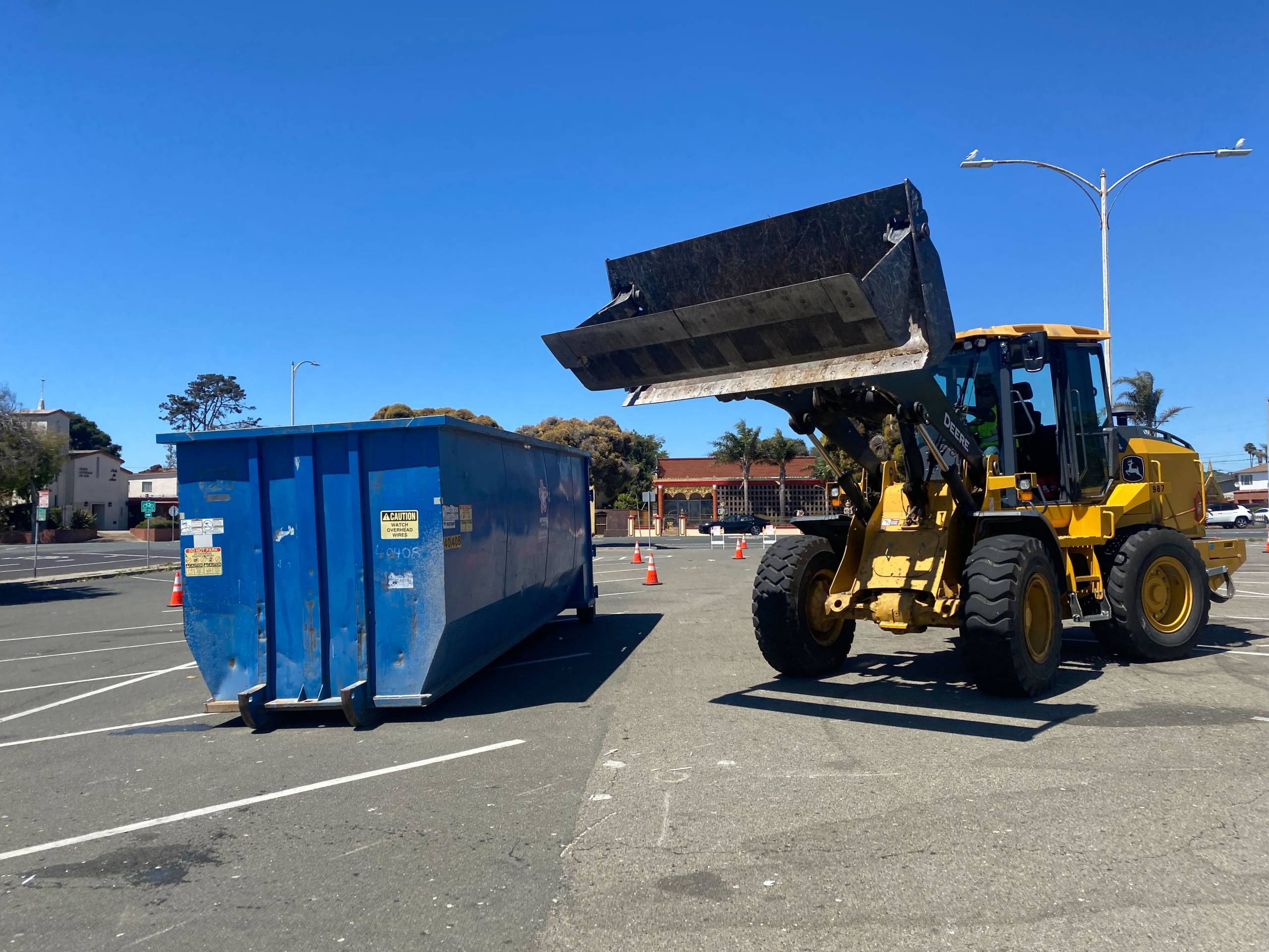 A person operates a construction vehicle next to a blue dumpster.