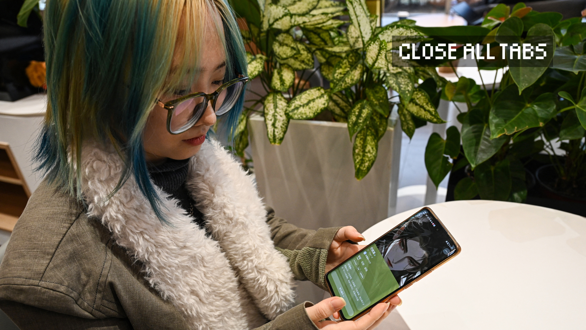 A young woman with glasses and multicolored hair sits at a café table, holding a smartphone. On her phone screen is an anime-style avatar from an AI chatbot app. She appears focused as she looks at the screen. The background includes potted plants and another seated person.