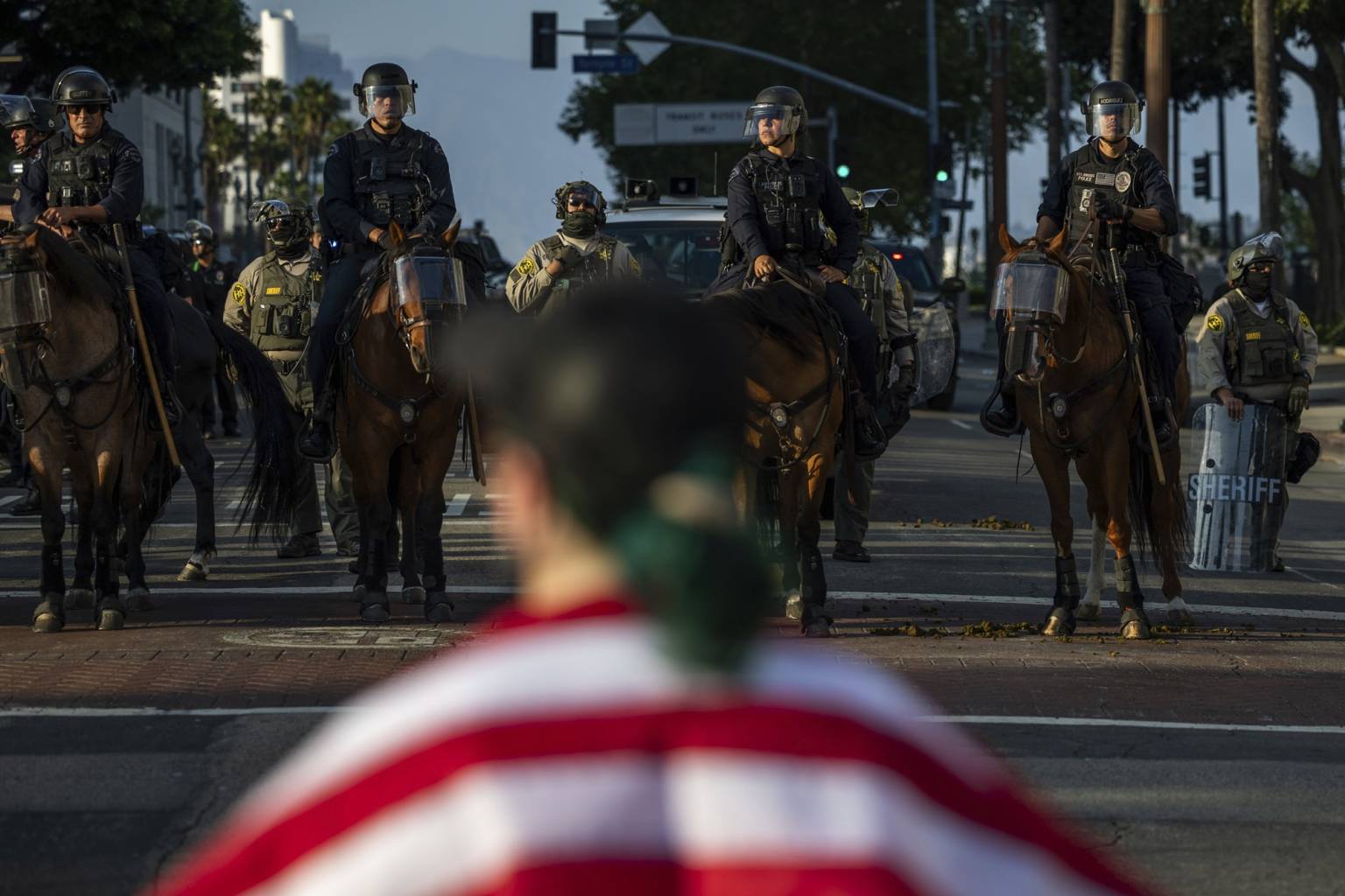 Una persona lleva puesta una bandera de Estados Unidos y enfrente de ella aparecen varios agentes de policía montados en caballo.