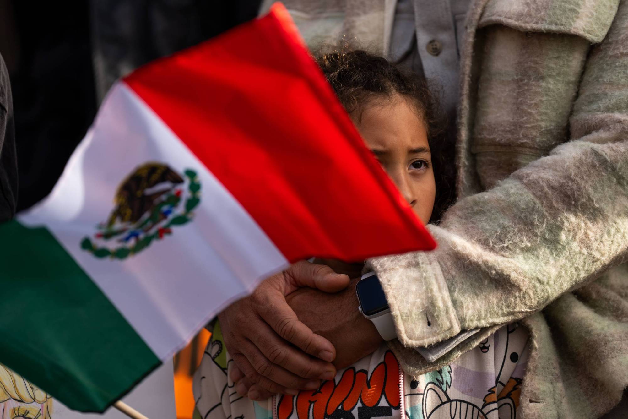 Un adulto abraza a una niña que lleva en la mano una bandera mexicana.