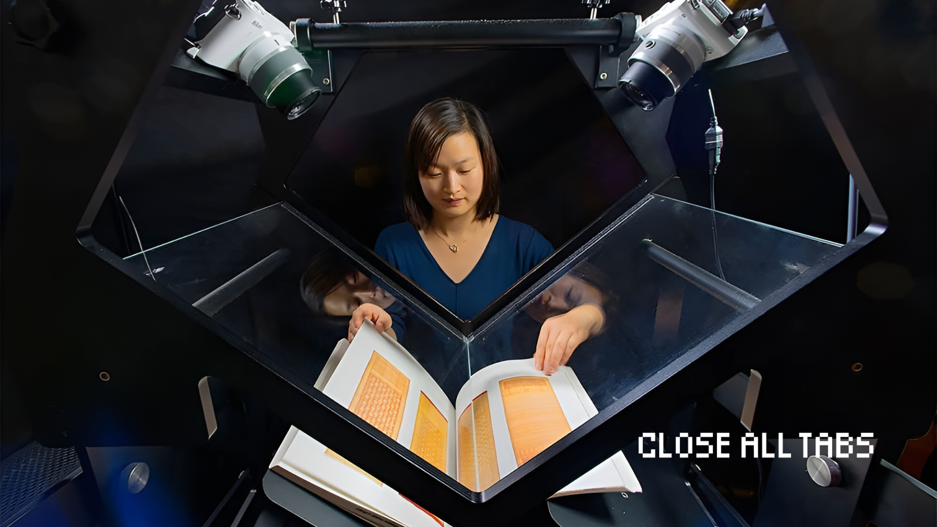 A scanning technician at the Internet Archive sits at a digitization station, carefully turning the pages of a large manuscript under angled glass. Above her, two digital cameras are mounted to capture high-resolution images of each page as she turns them by hand. The glass enclosure helps keep the pages flat while protecting the material. The Close All Tabs logo appears in pixelated font in the lower right corner.