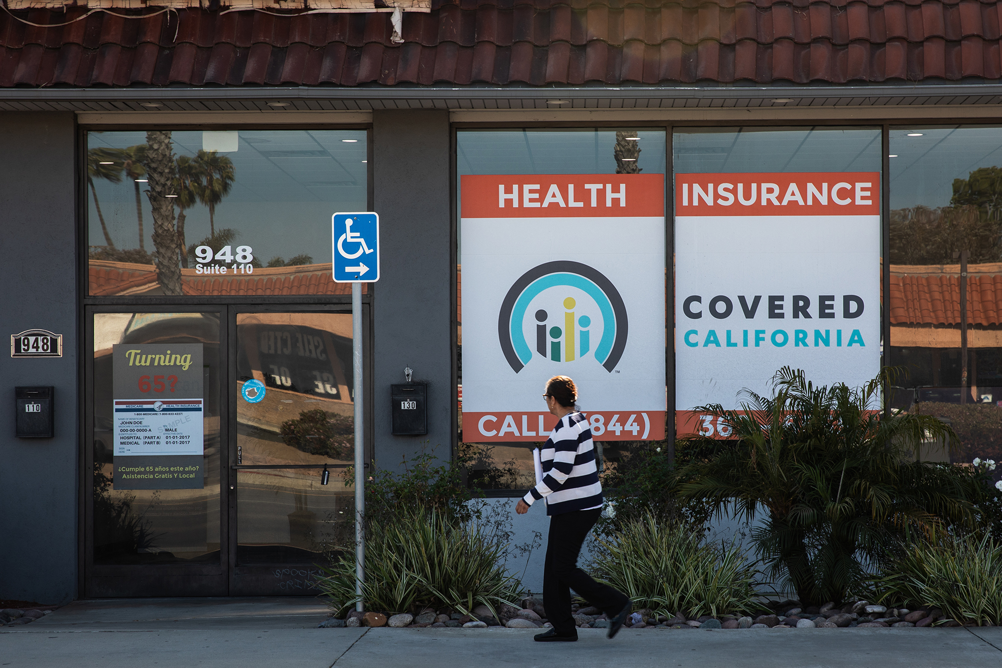 A woman wearing a black and white striped shirt walks past a building with a sign that says "Health Insurance Covered California."