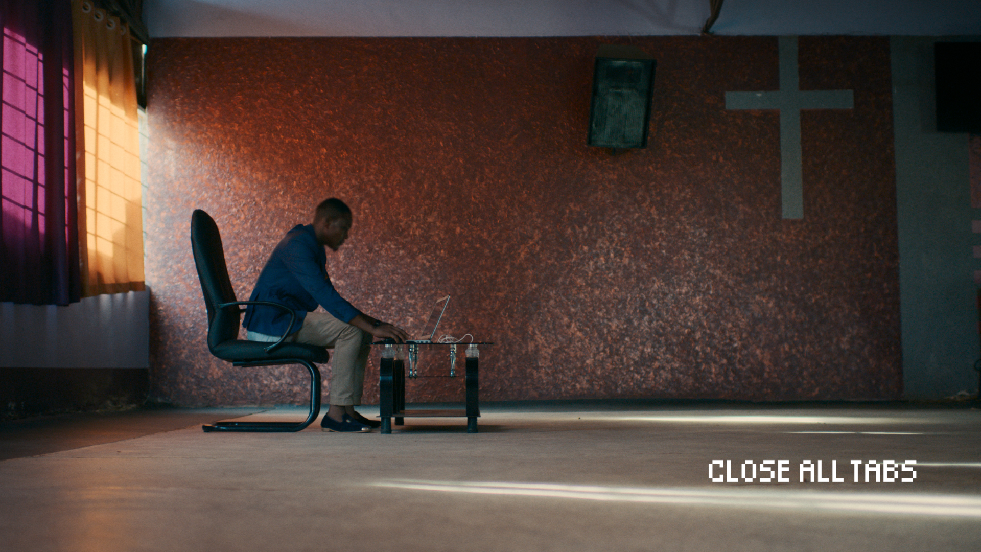 A wide image of a Kenyan man sitting in a desk chair on his laptop in a mostly empty room. The laptop is placed on a glass coffee table in front of him. Orange light streams in from curtained windows behind him to the left of the screen. A large cross hangs on a brown and gold wall behind him. The floor is bare and dappled in light. The Close All Tabs logo appears in pixelated font in the lower right corner.