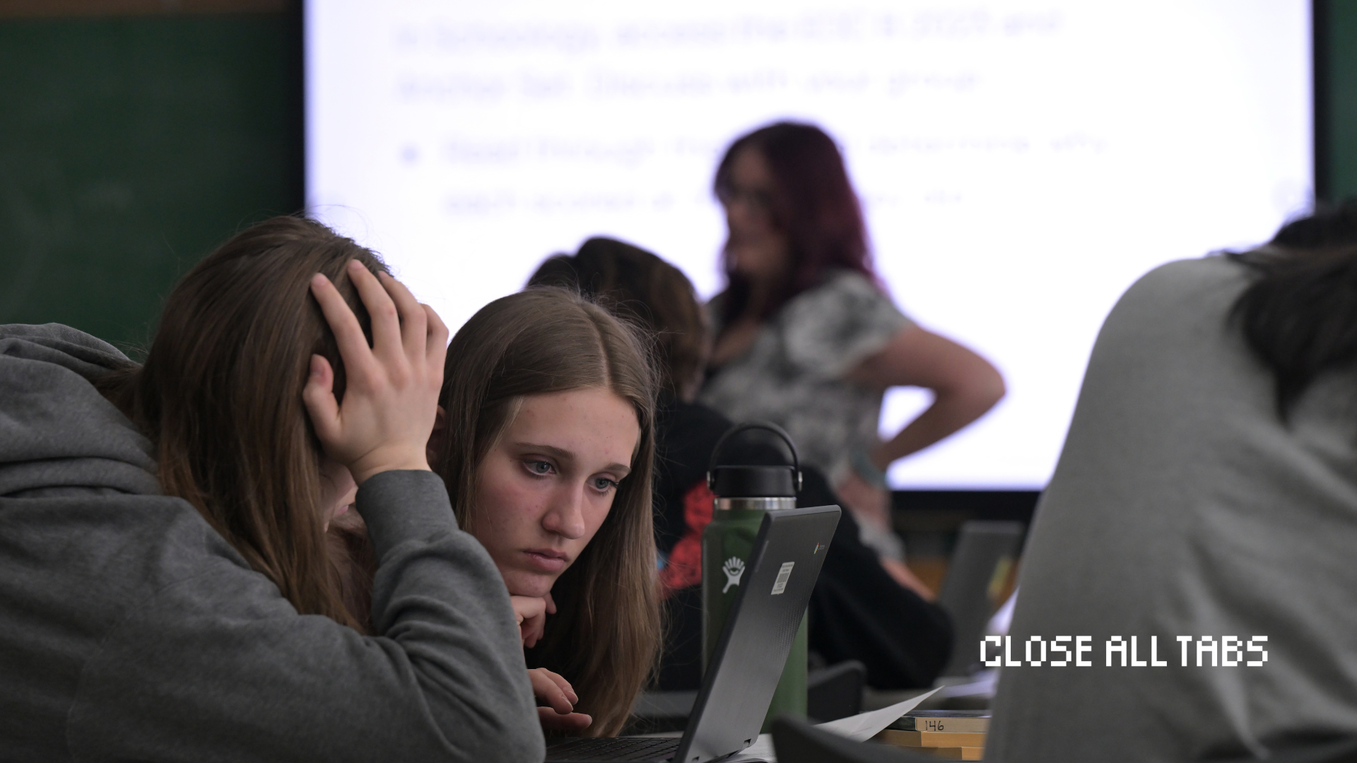 Two female high school students hover over a computer. One student has a hand covering her face as the other student leans in closely to the computer screen.Their teacher and a projection of the lesson are blurred in the background. The Close All Tabs logo appears in pixelated font in the lower right corner.