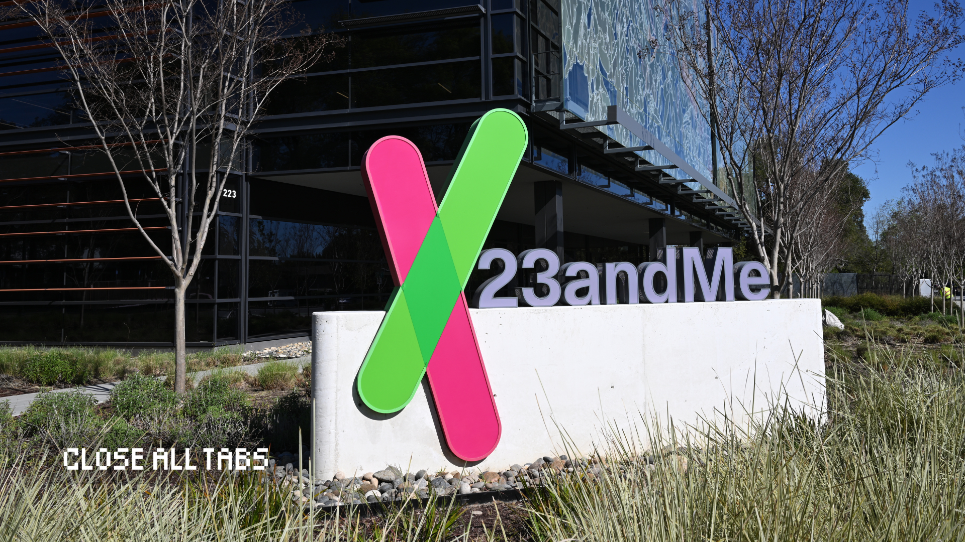 An image of the 23andMe headquarters in Sunnyvale, California. The company’s sign is prominently displayed — it contains the 23andMe logo in lavender font with a pink and green X next to it. The sign is on top of a white stone wall in the grass in front of a textured glass building. The Close All Tabs logo appears in pixelated font in the lower left corner.