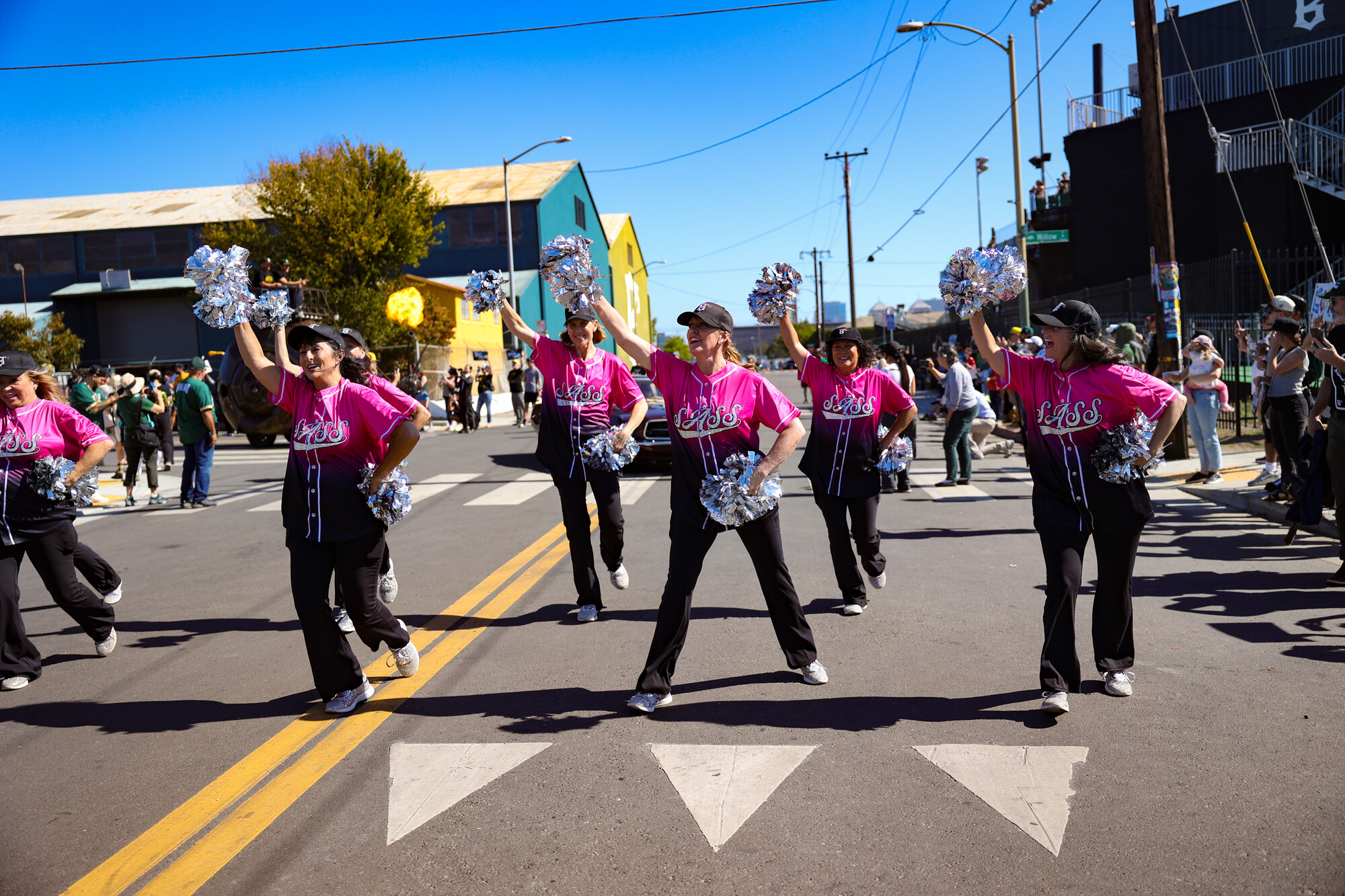 Six people wearing pink shirts and black pants dance and wave pom poms in the street.