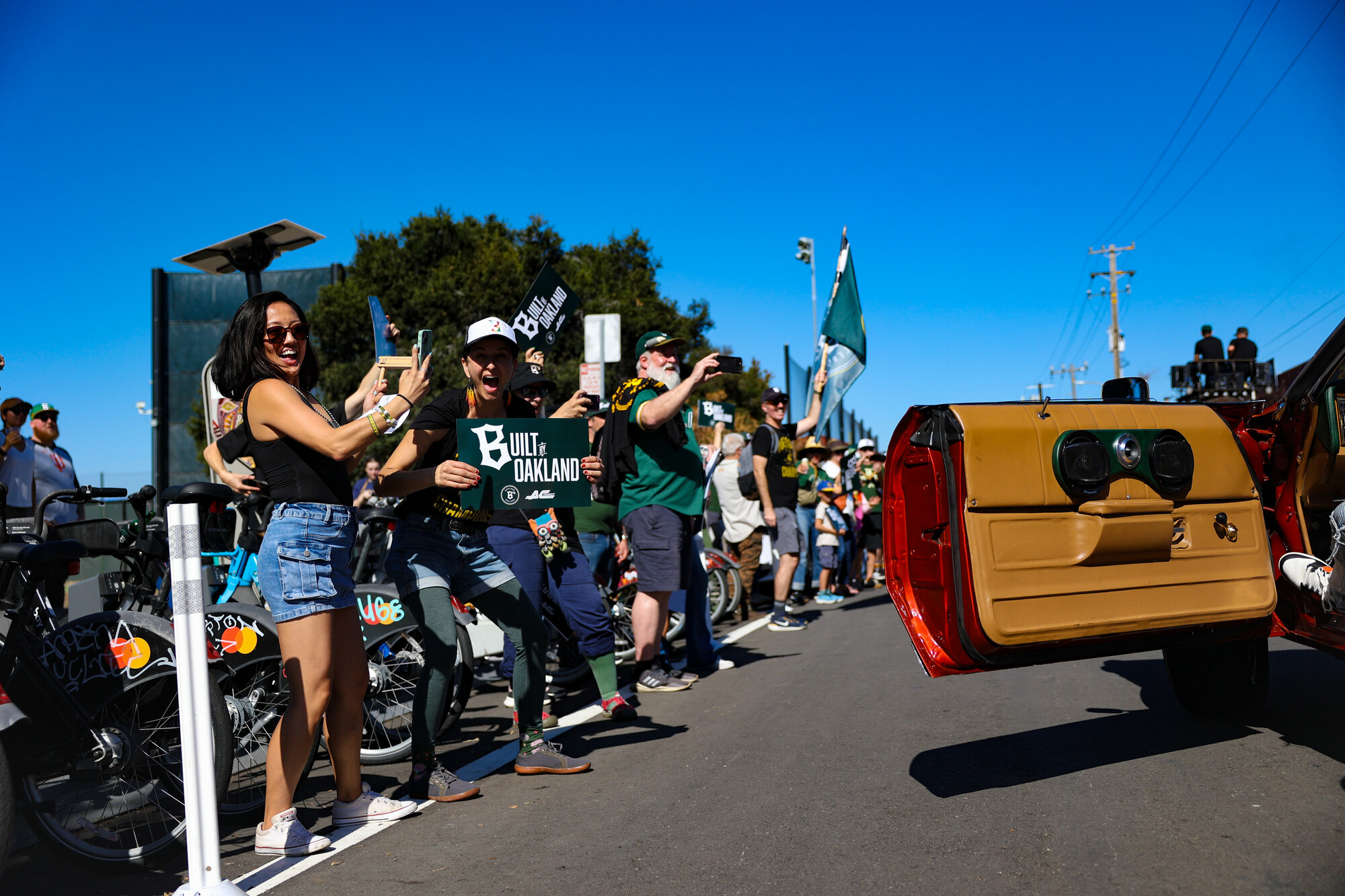 People on the side of the street wave, hold signs and take photos of people in a red car.