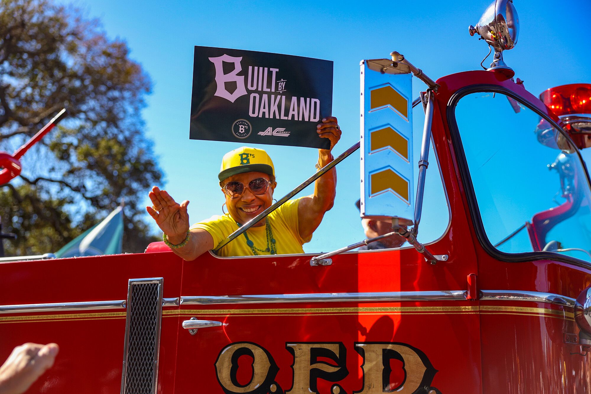 A Black woman wearing yellow clothing waves while holding a green sign that says "Built by Oakland" in a red vehicle.