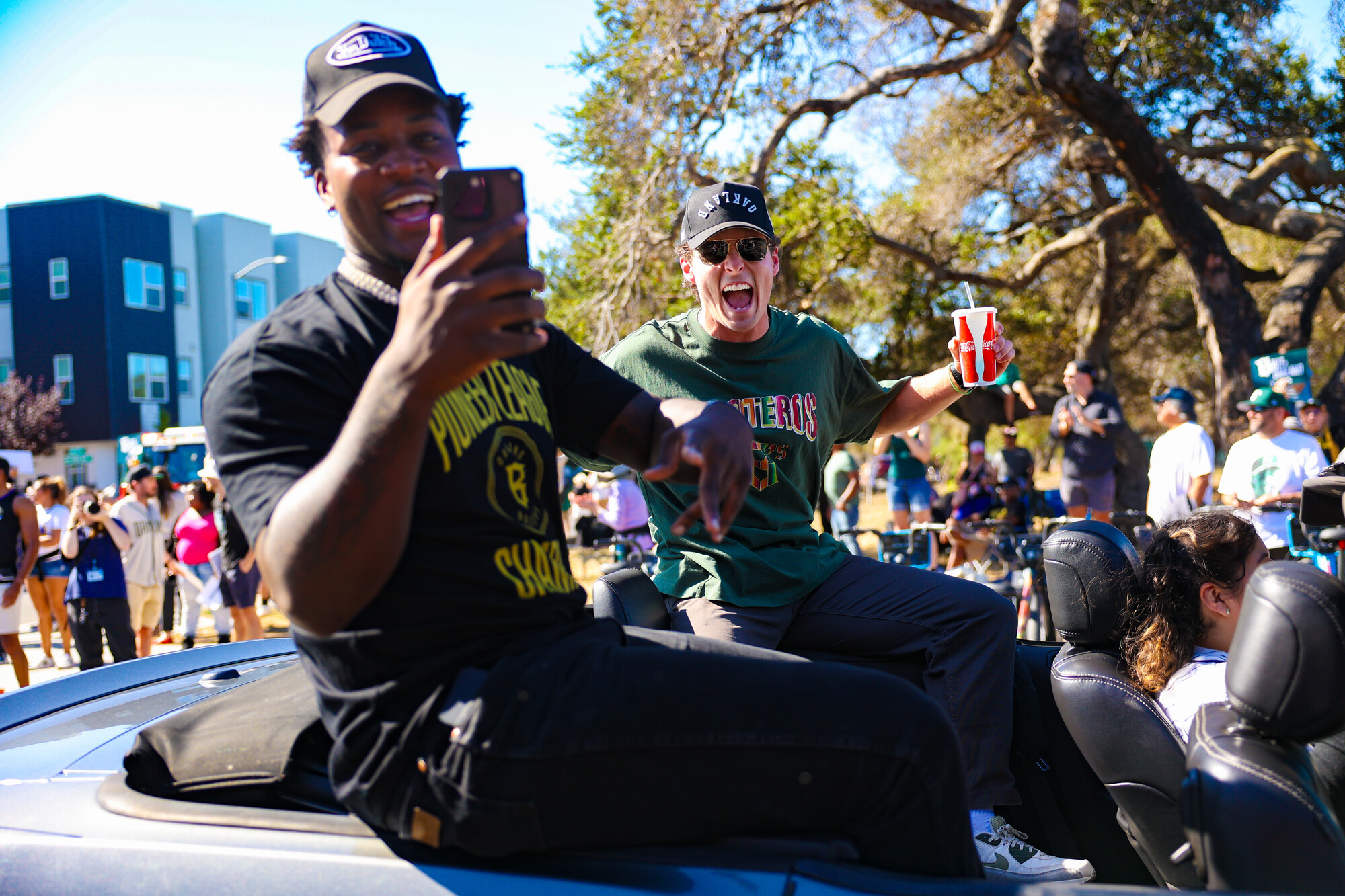 A Black man and white man pose while seated in the back of a topless car.