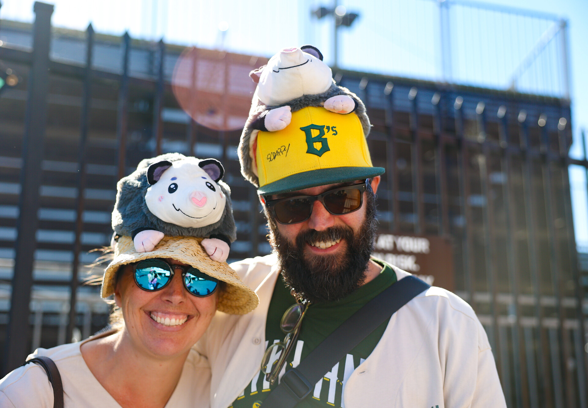 A man and woman pose together. They both are wearing hats with a toy possum on top as well as sun glasses.