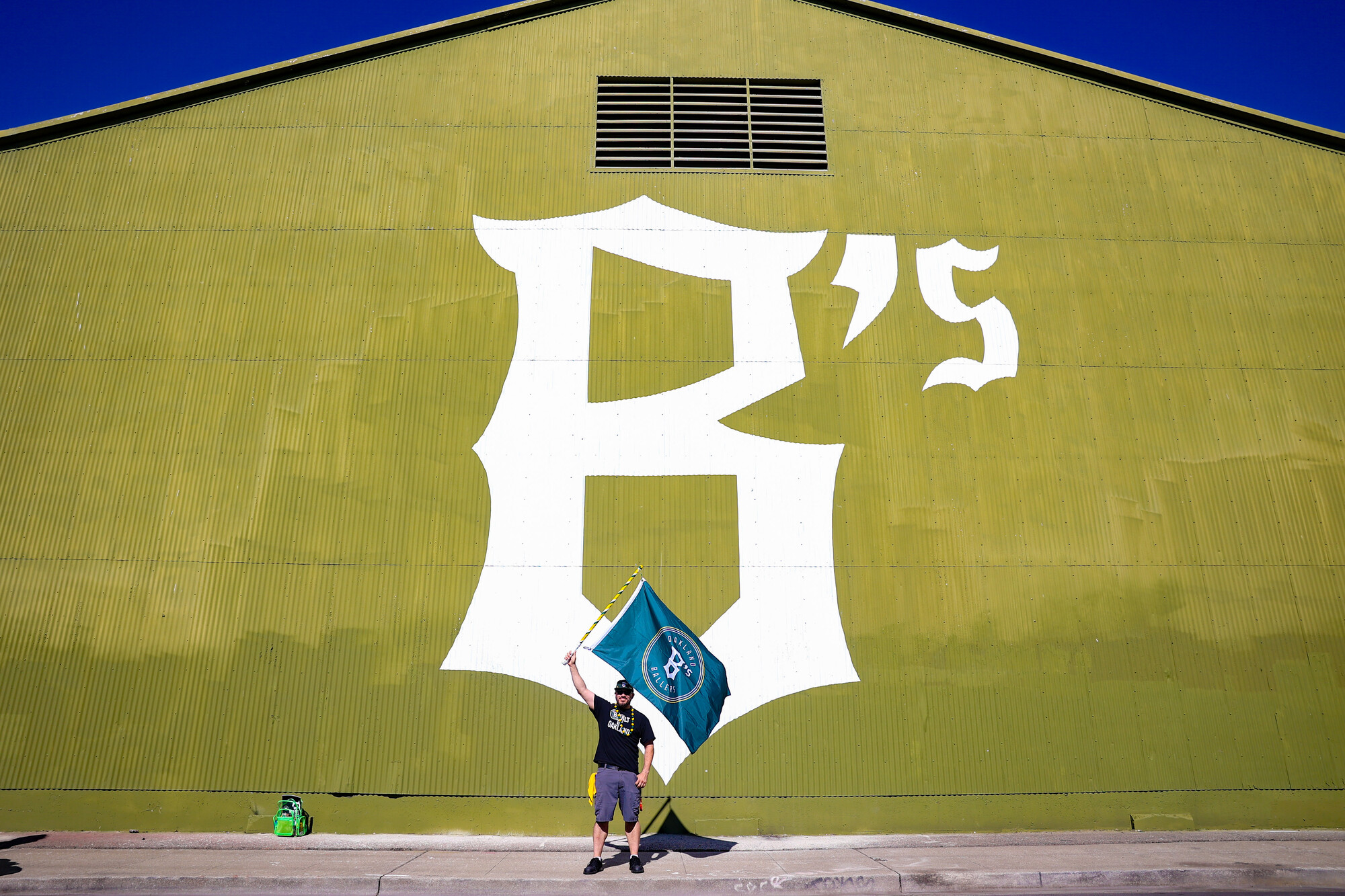 A man holding a green flag stands in front of a building with "B's" painted in white.