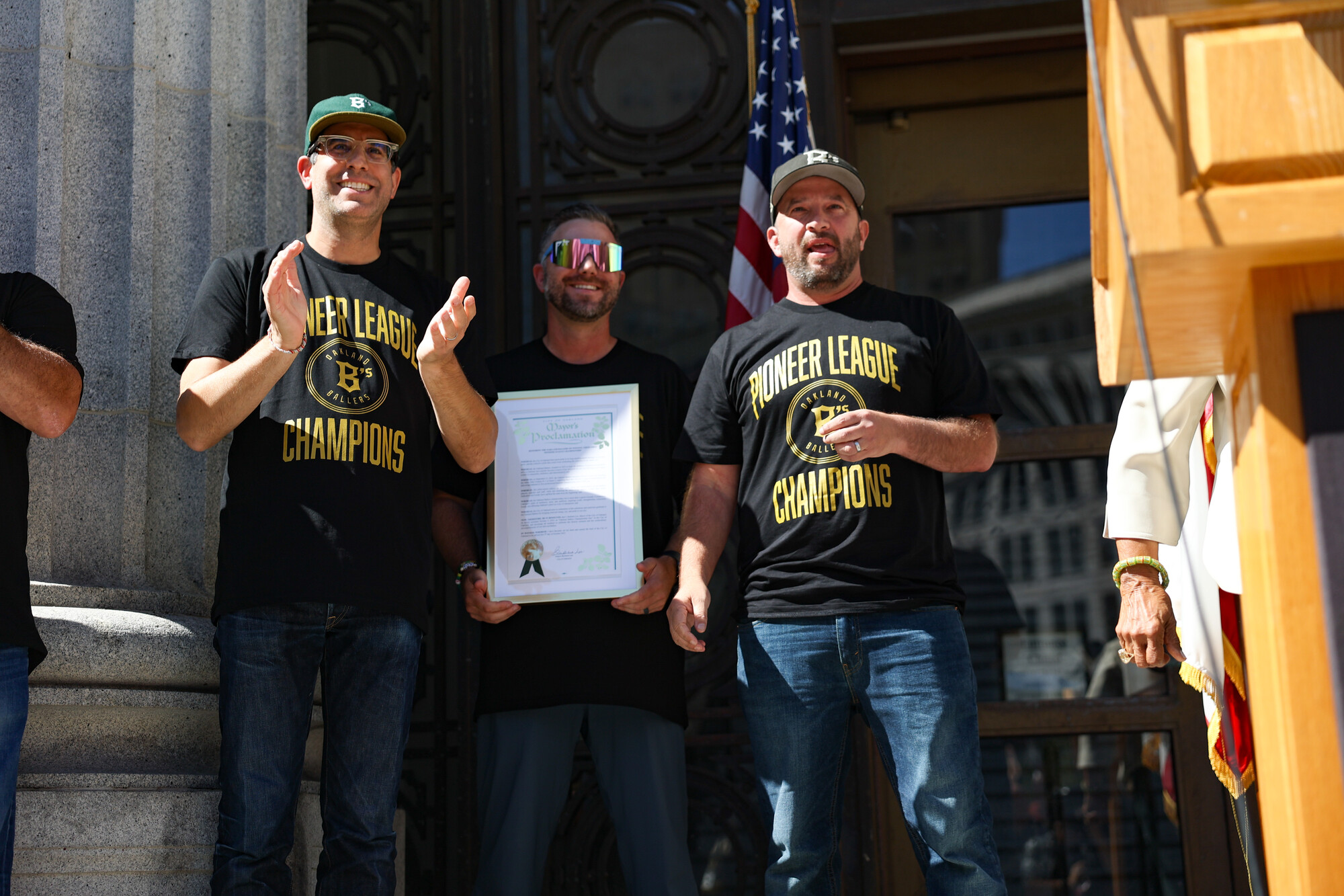 Three men wearing t shirts stand on the front steps of a building. The man in the middle is holding a large plaque.