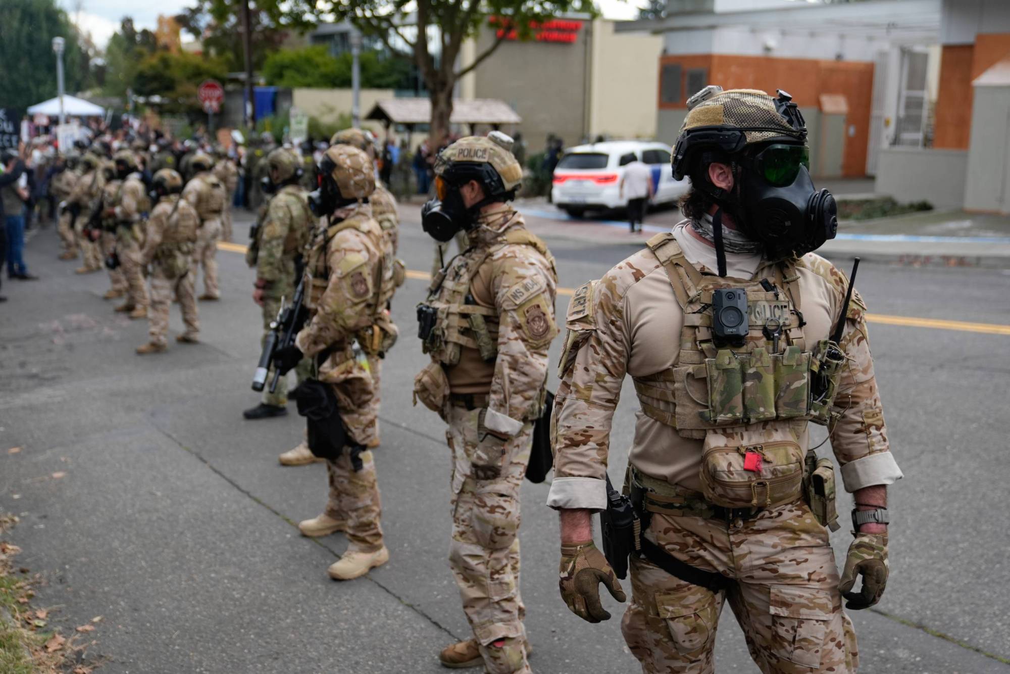 Several people dressed in military gear and wearing gas masks stand in a line in the middle of the street.