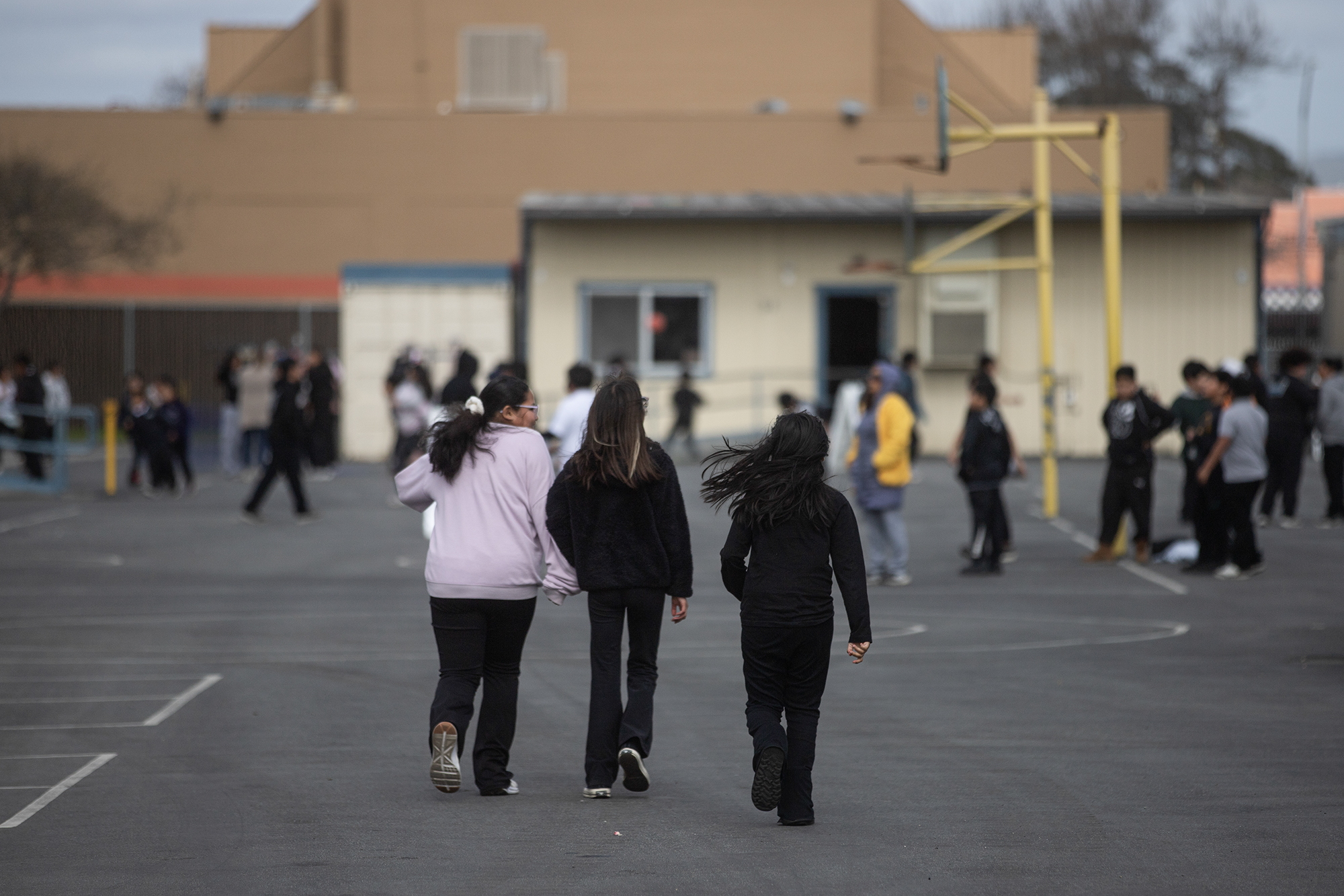 Several children on a playground and basketball court with three facing away from the camera.