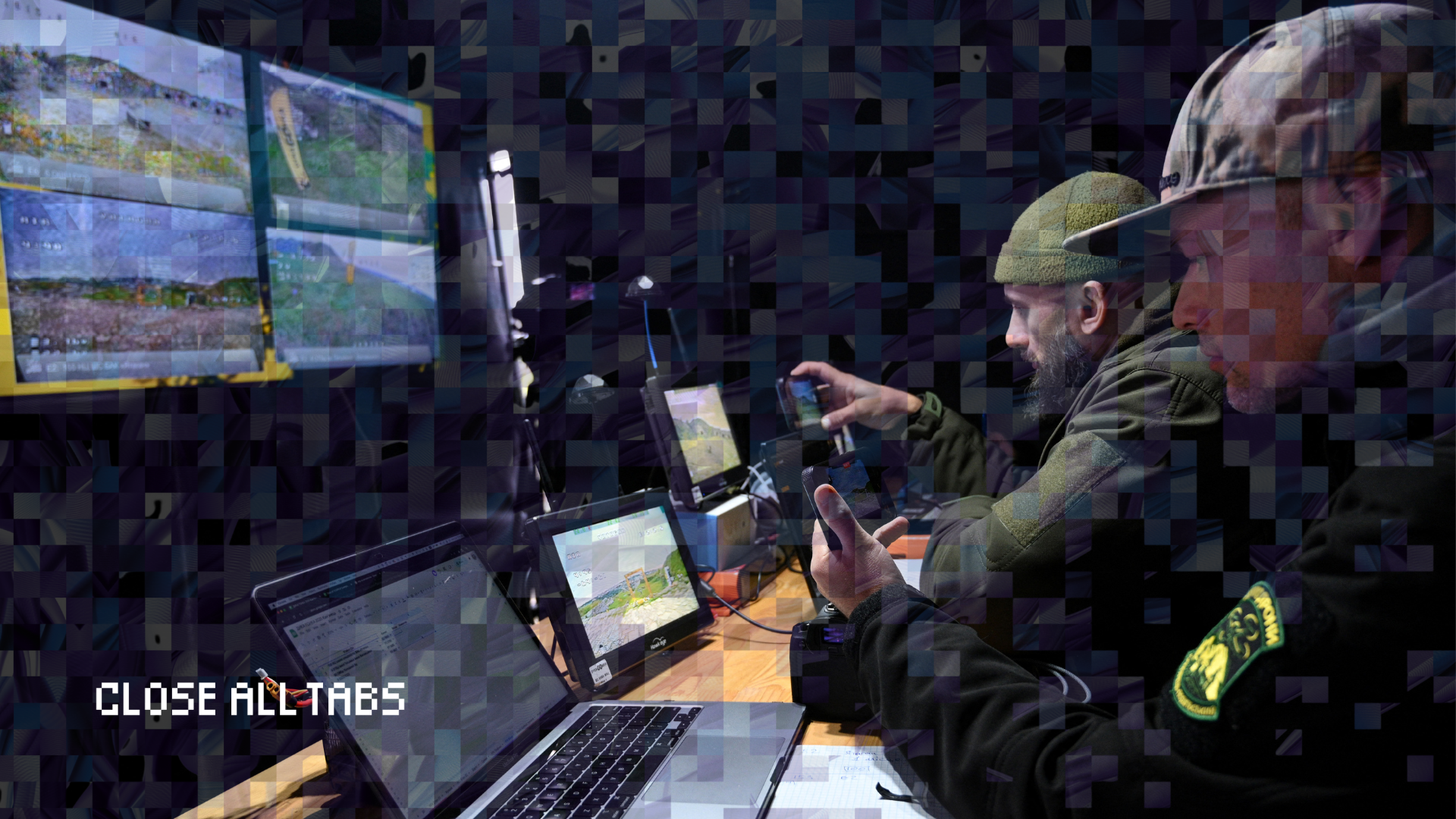 Two Ukrainian service members sit inside a dimly lit mobile command center, monitoring several screens showing drone footage. Laptops, monitors, and handheld devices cover the desk in front of them. A pixelated overlay obscures parts of the image, and the ‘Close All Tabs’ logo appears in the lower left corner.