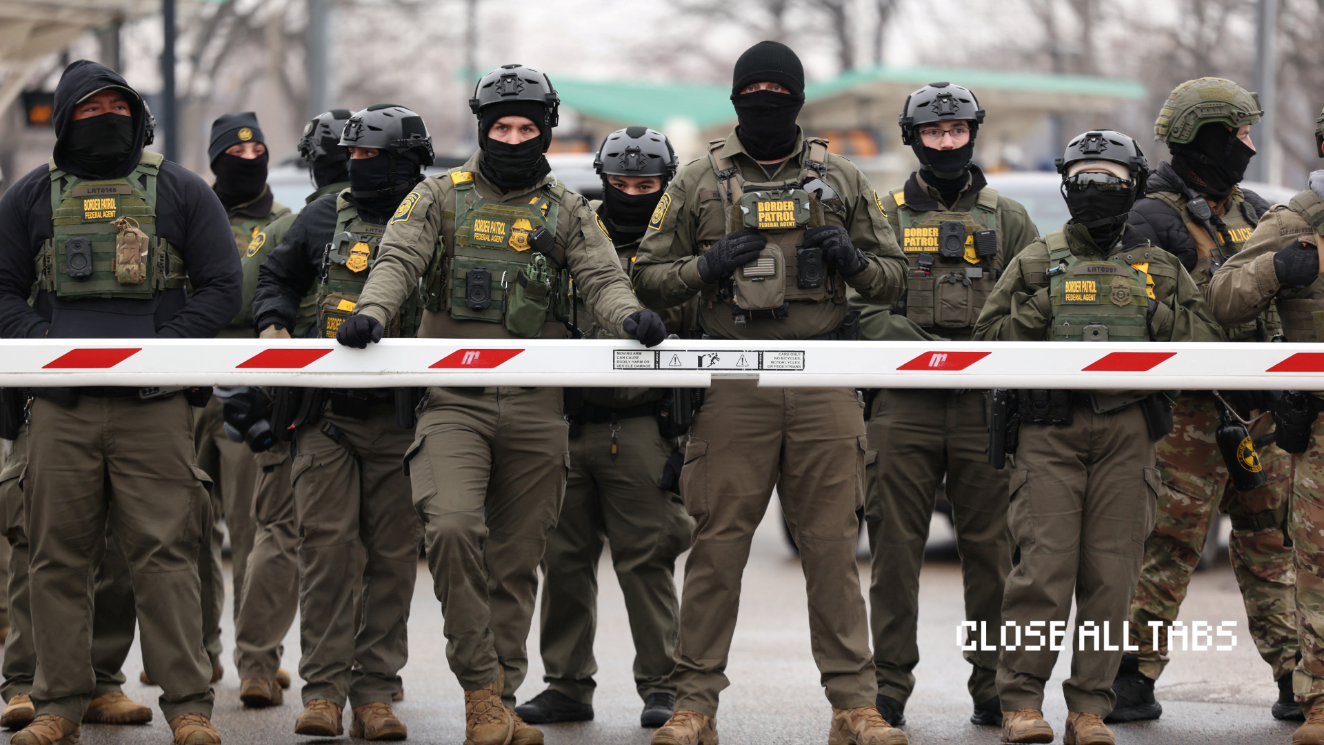 A line of U.S. Border Patrol agents wearing helmets, tactical vests, and face coverings stand shoulder to shoulder behind a metal crowd-control barrier, obscuring their identities, as they block a street during a law enforcement operation.