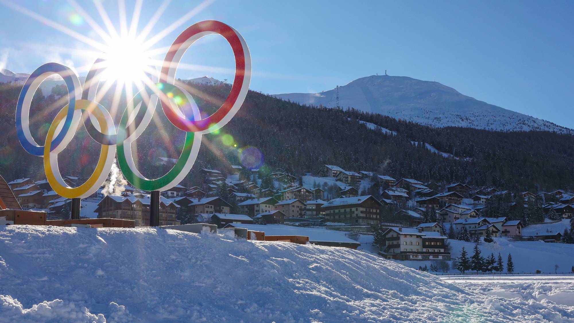 The sun shines through the Olympic rings with mountains and a village behind.