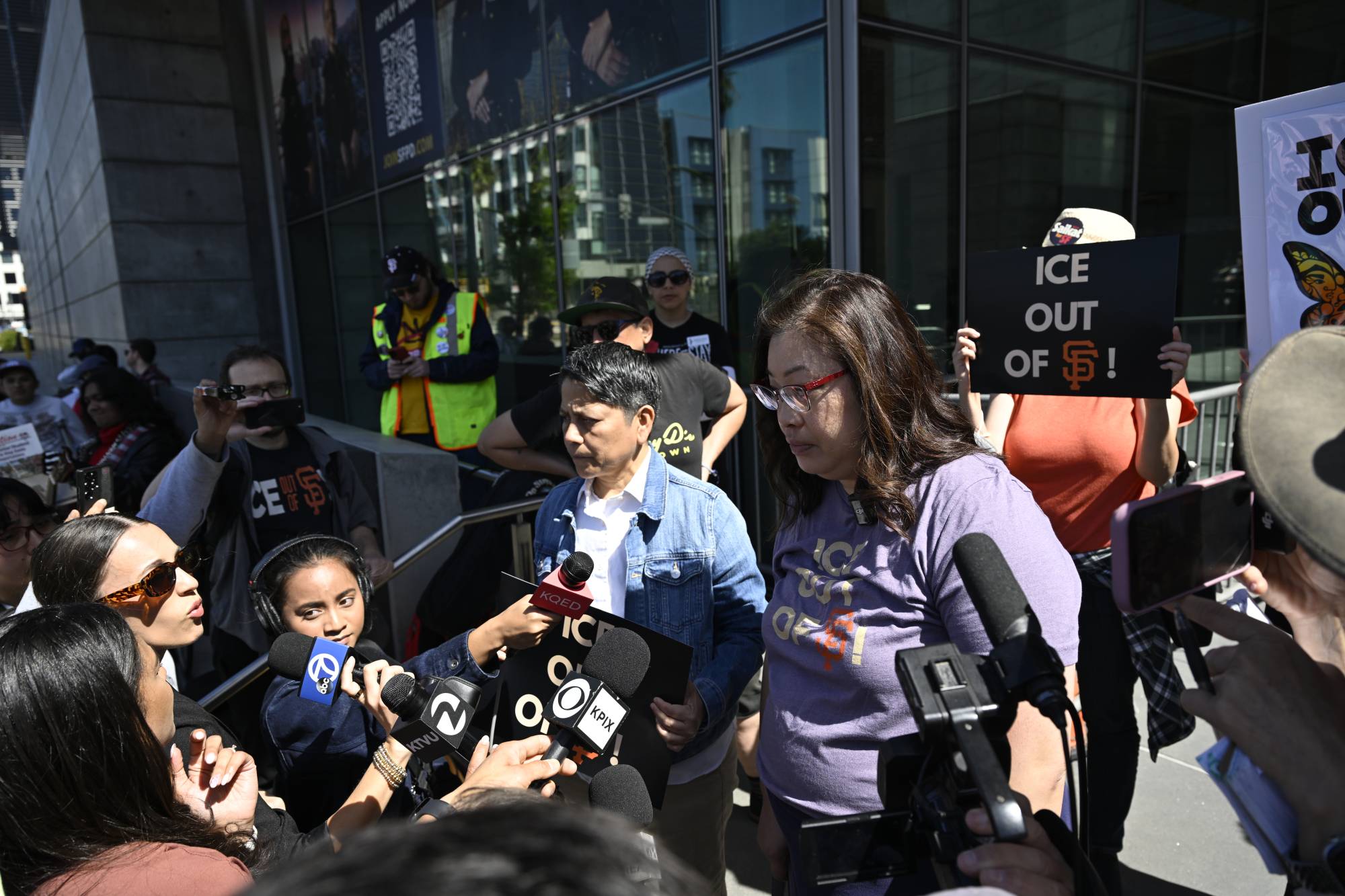 Two people speak into news microphones as a crowd of protesters surround them. A sign says "ICE out of SF"