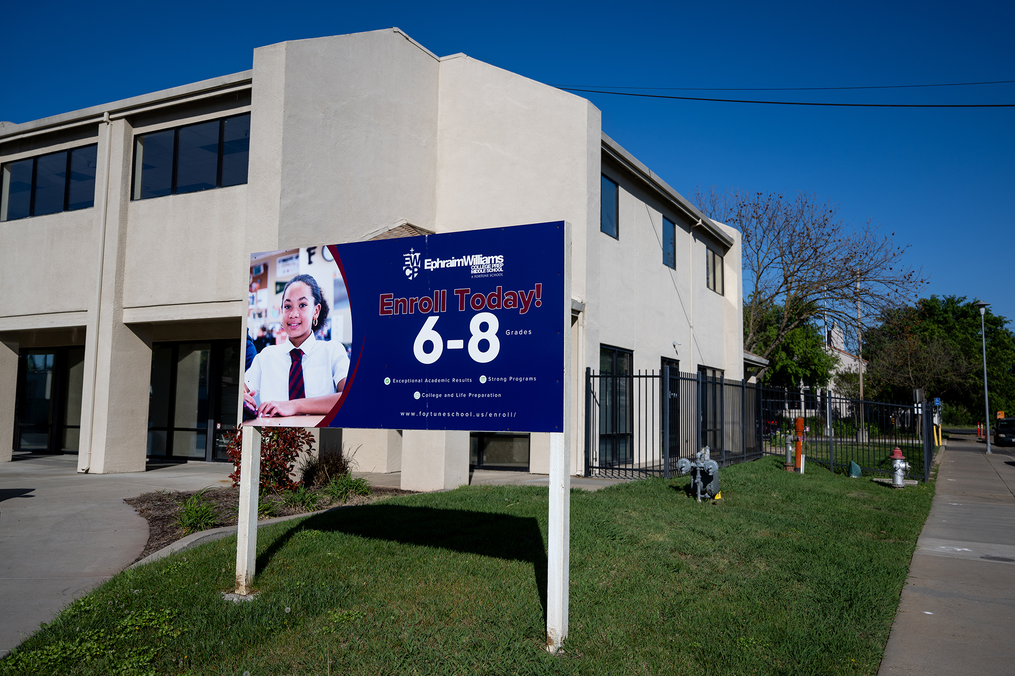 A school building with a sign in front of it that shows a photograph of a student and text that reads, &ldquo;Enroll Today! 6-8 grades.&rdquo;