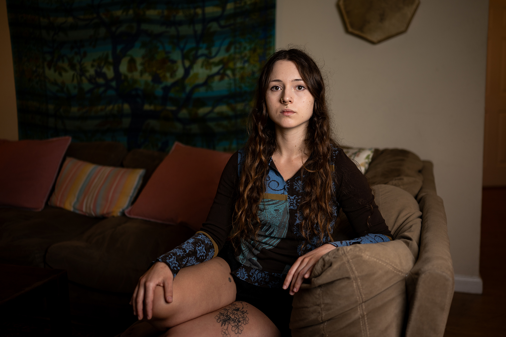 A woman in her twenties sits on a sofa and looks at the camera with a serious expression.