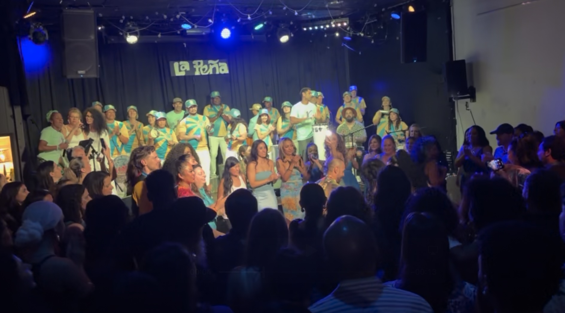 A lively crowd gathers around a dance floor inside a small music venue as a large percussion ensemble performs on stage beneath blue lights. Audience members clap and dance close to the performers, creating an energetic, celebratory atmosphere. A sign reading “La Peña” hangs above the stage.