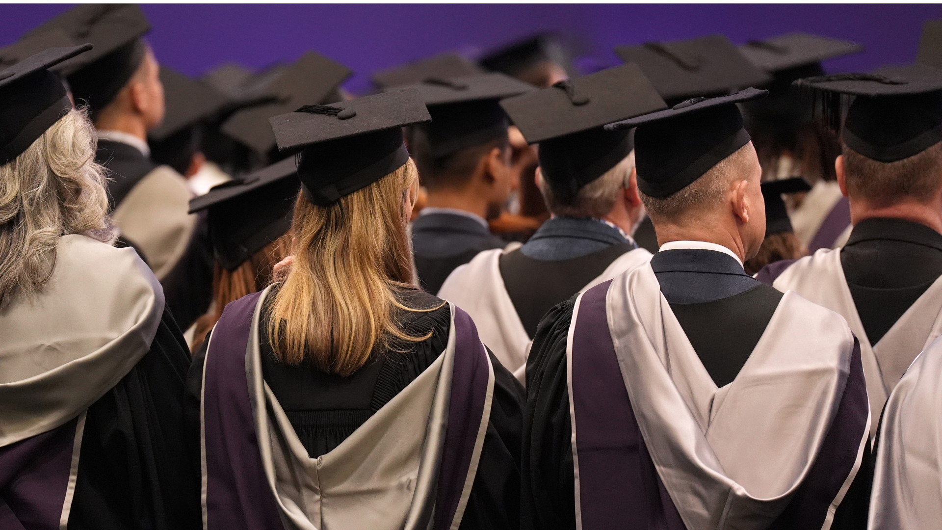 Graduates during a graduation ceremony. Graduates have their back turned away from the camera and are wearing black graduation caps and black and purple gowns.