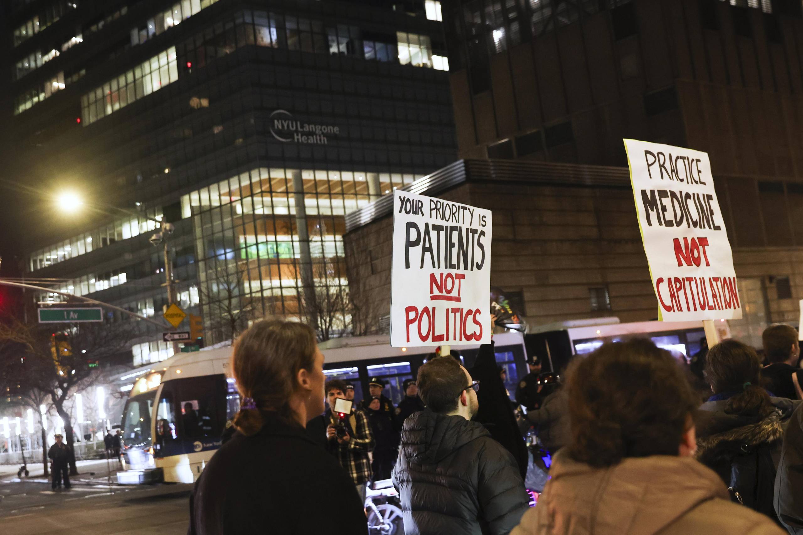 People are seen carrying two signs during a nighttime protest. One says, "Your priority is patients, not politics." The other says, "Practice medicine, not capitulation." The logo "NYU Langone Health" is displayed one of the buildings. 