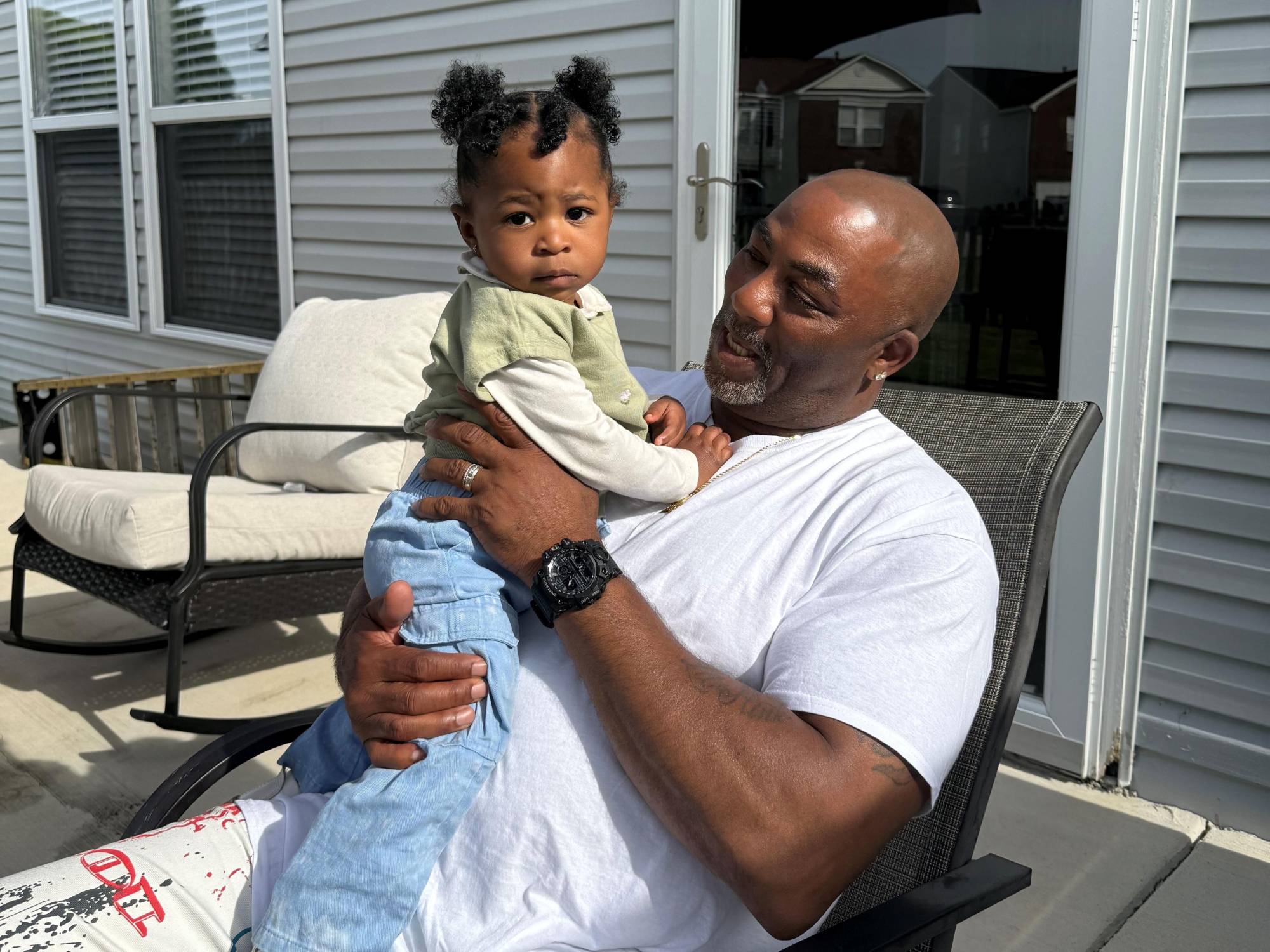 Anthony Bailey, 61, sits on a chair on a porch outdoors while holding a baby grandchild.
