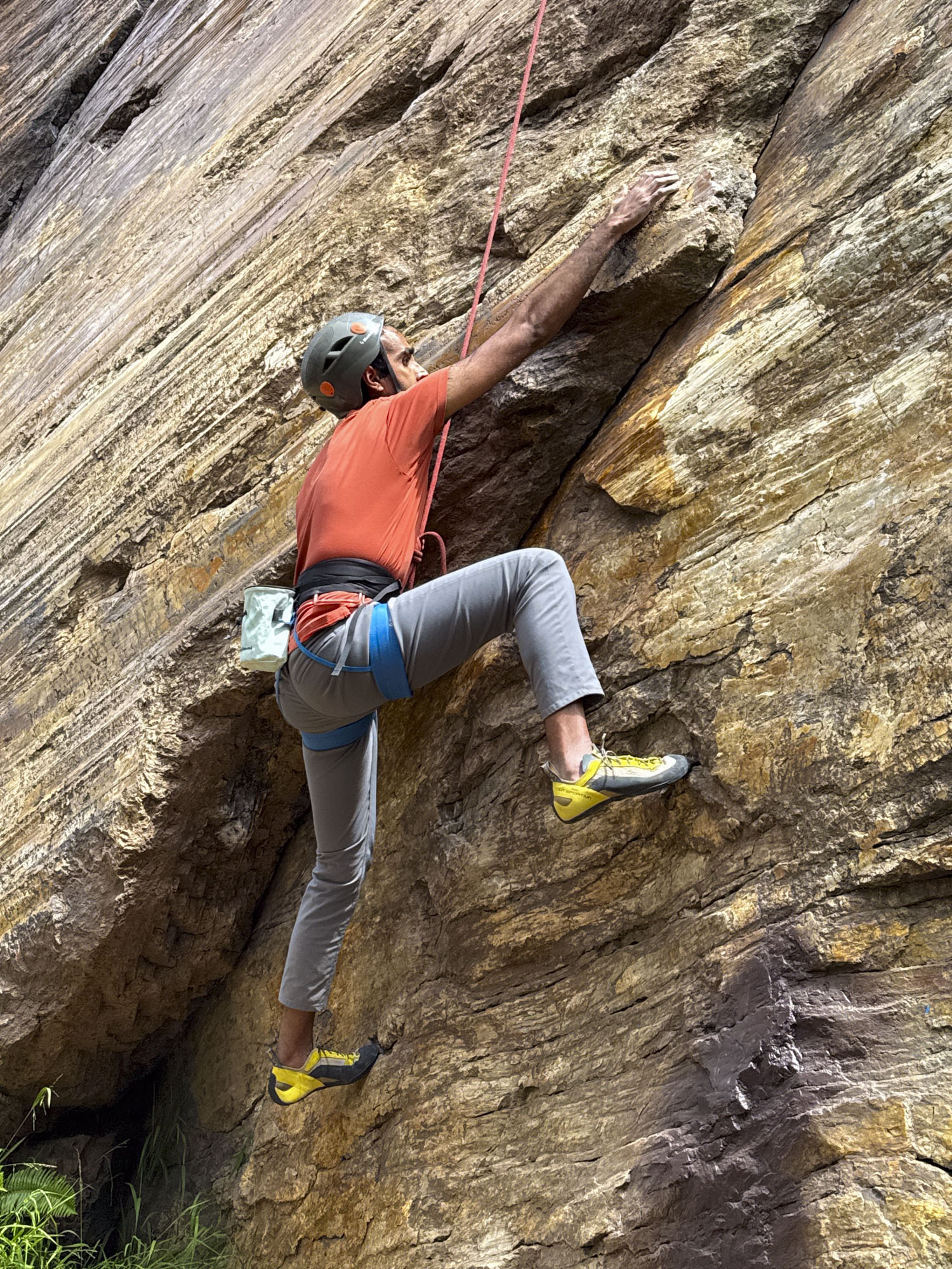 A man in orange shirt and grey pants wearing a helmet and rock climbing shoes climbs a rock wall.