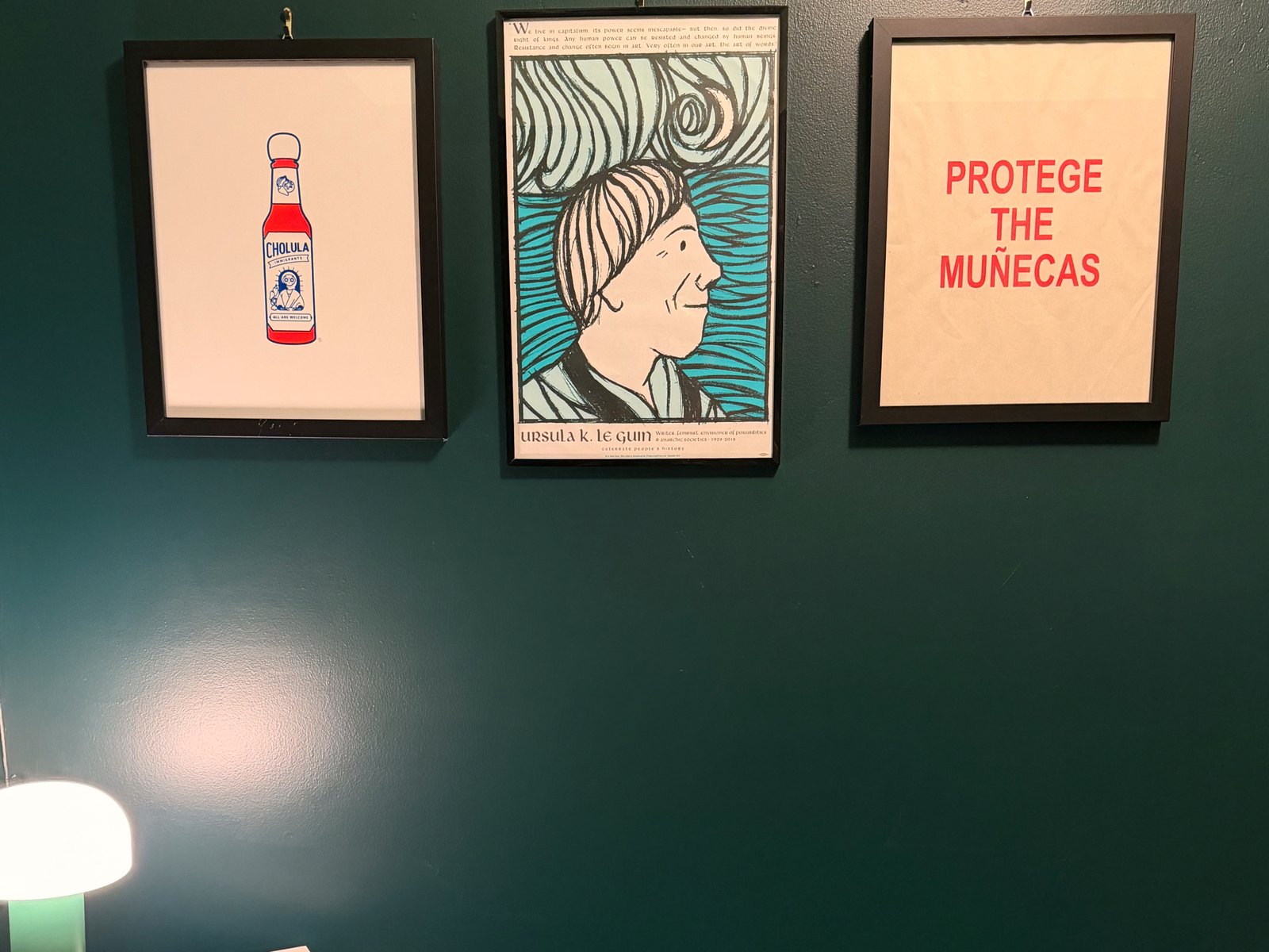 Three framed prints on a dark green wall: a hot sauce bottle labeled “Immigrants — All Are Welcome,” an Ursula K. Le Guin “Celebrate People’s History” poster, and a print reading “Protege the Muñecas.”