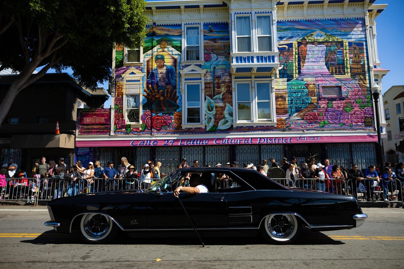 A black lowrider cruises past a crowd in front of a muraled Victorian in the Calle 24 Latino Cultural District.