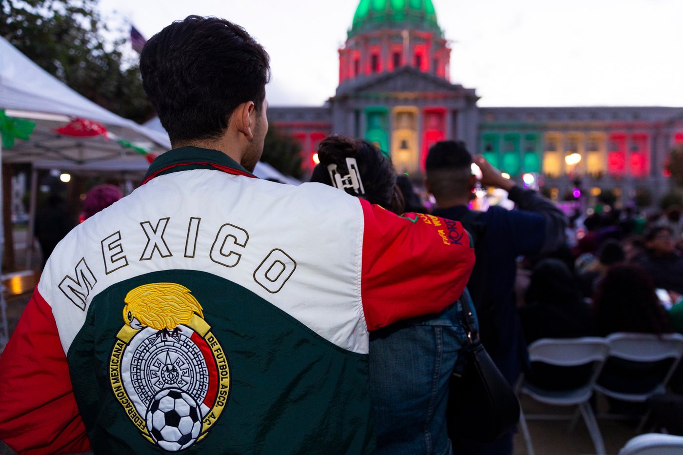 A man in a Mexico national team jacket faces San Francisco City Hall, lit in red, white and green, during an El Grito celebration.