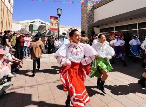 Ballet folklórico dancers in red and green skirts perform in an outdoor shopping plaza.