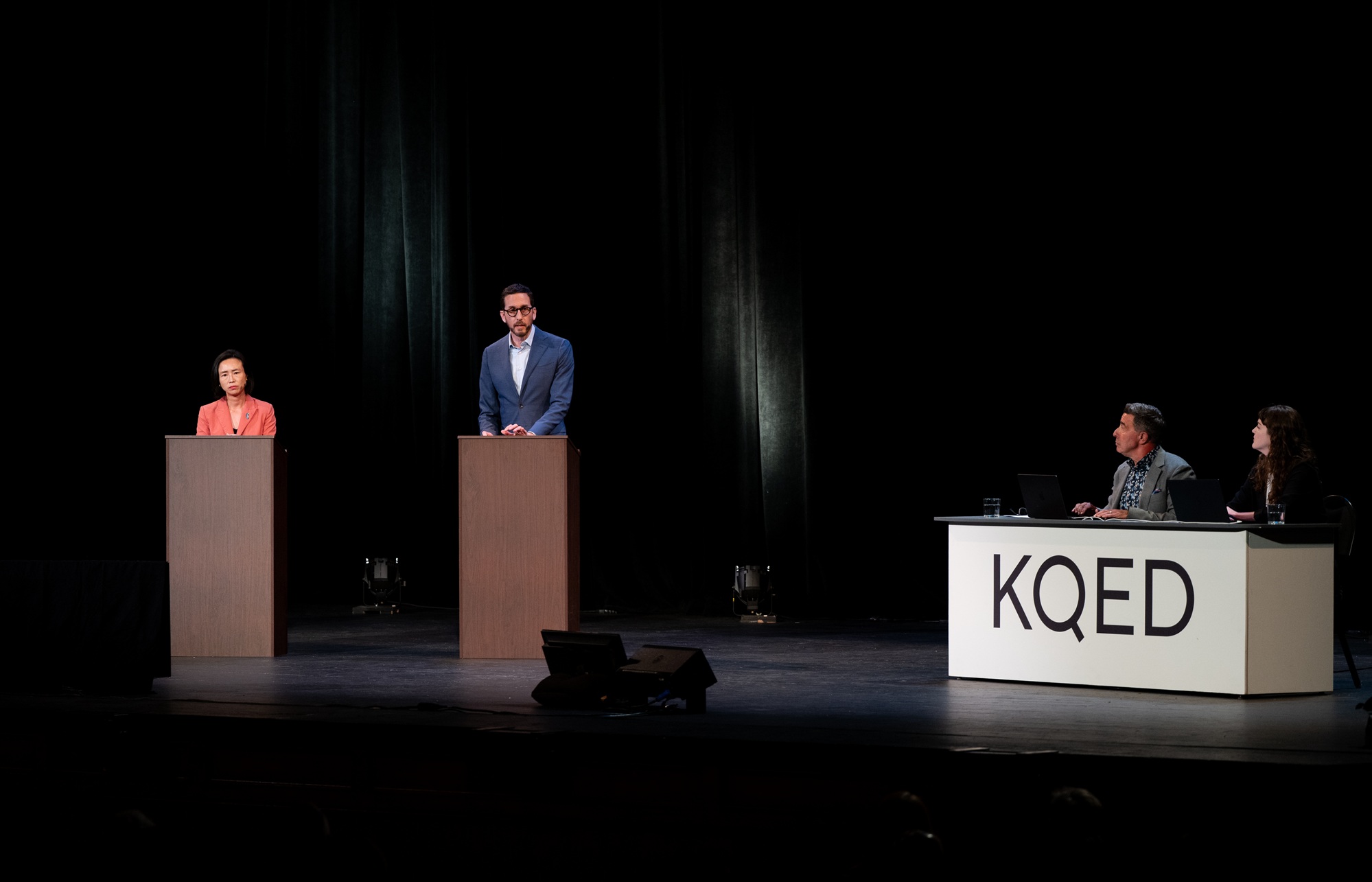 Two people stand at lecterns on a stage while two moderators sit nearby at a desk