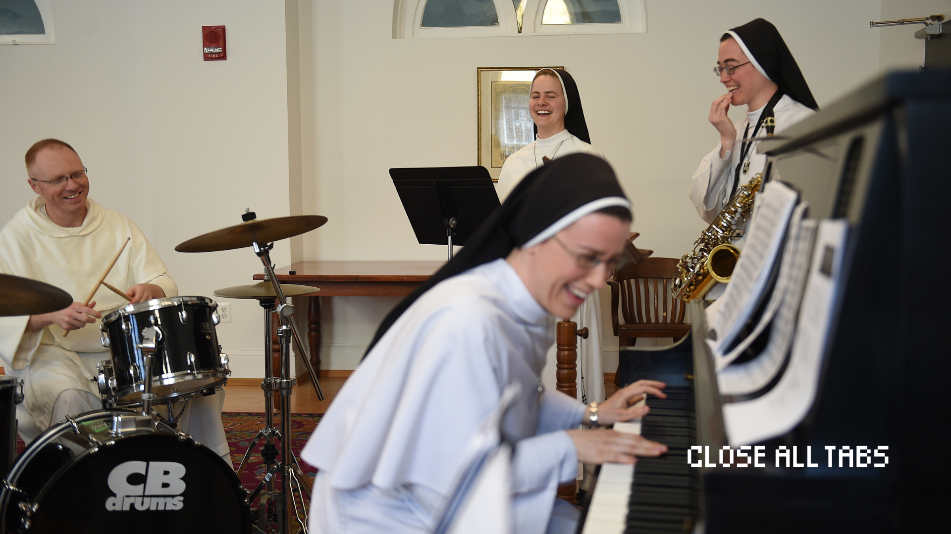 Image of the inside of what seems to be a church. The right half of the image, in the foreground and slightly blurry, is a nun with glasses, sitting at a black piano and laughing. Her hands lay on the keys and there is sheet music in front of her. Behind her, on the left of the image, is a man in religious dress, sitting behind a black drum set, also laughing. To the right, two Catholic sisters are standing, also laughing. The one closest to the man is behind a black music stand and the furthest from him is carrying a saxophone around her neck. The text "Close All Tabs" appears in white text in the lower right corner.