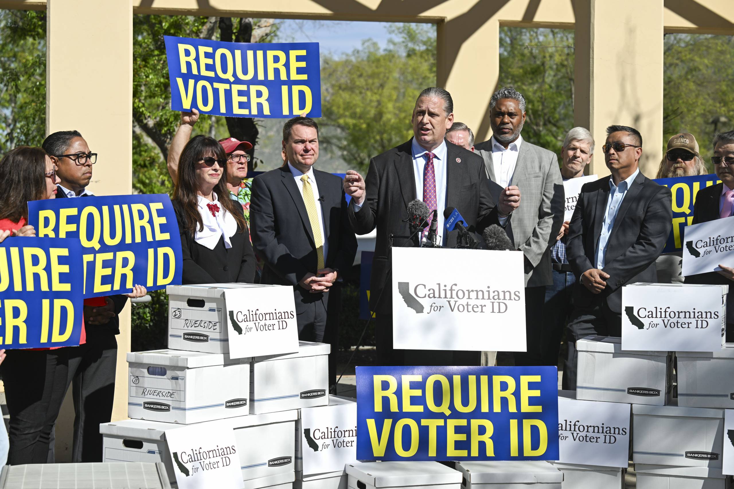 People in suits present at a podium surrounded by signs that read "Require Voter ID."