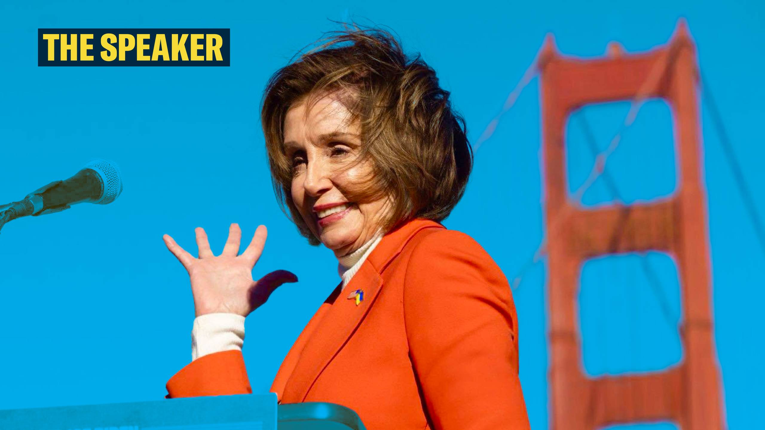 Nancy Pelosi stands behind a podium, in front of the Golden Gate Bridge. She’s wearing a red suit and her right hand is gesturing toward the bridge.
