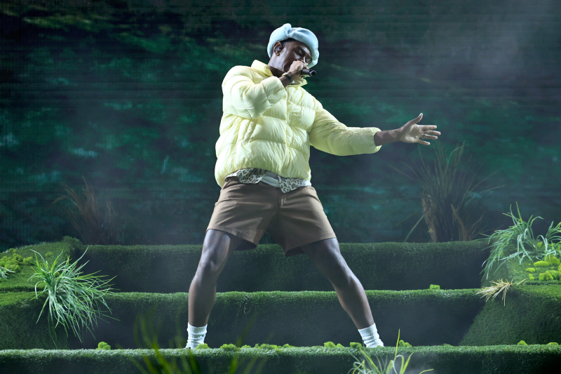 Tyler, the Creator performing onstage against a green backdrop wearing brown shorts and a lime green puffer jacket.