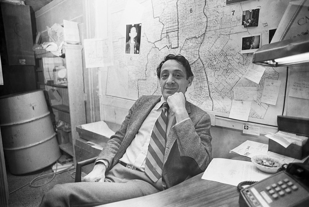 A white man in a suit and tie leans back in his chair behind a desk in his office and smiles at the camera.