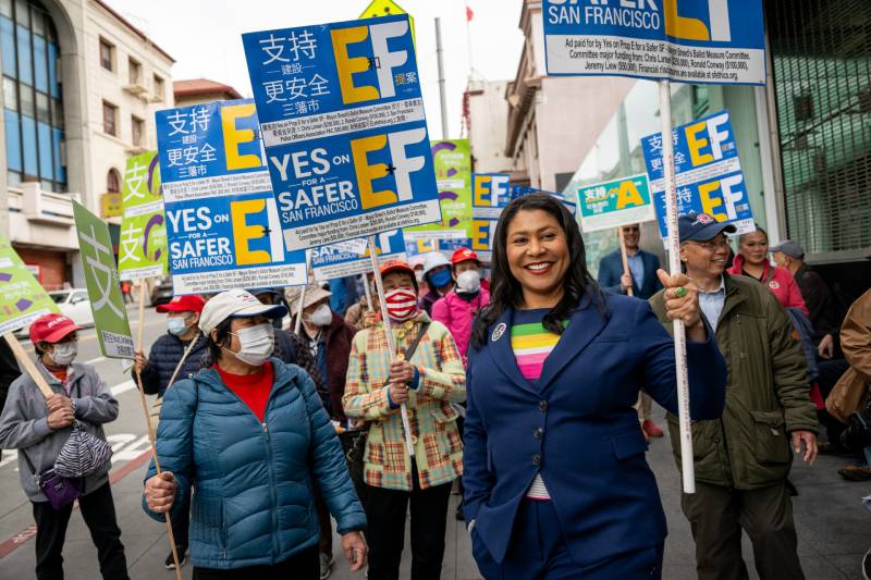 Mayor London Breed, in a blue suit and colorful shirt, smiles big, carrying a blue sign that says E and F with big letters.