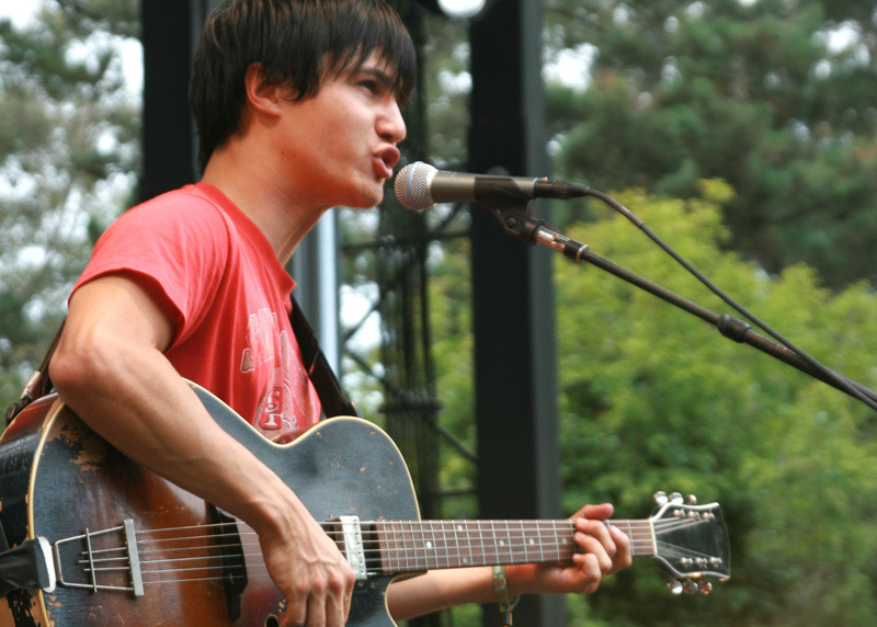 A man wearing a red t-shirt stands on a stage in the woods singing into a microphone and playing a guitar.